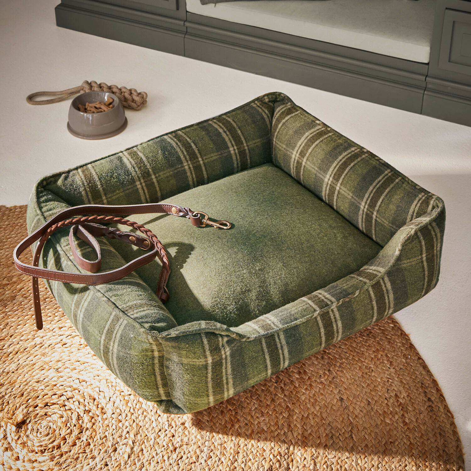 Zielone legowisko dla psa z miękkiego materiału w kratę z brązową skórzaną smyczą i miską na przysmaki