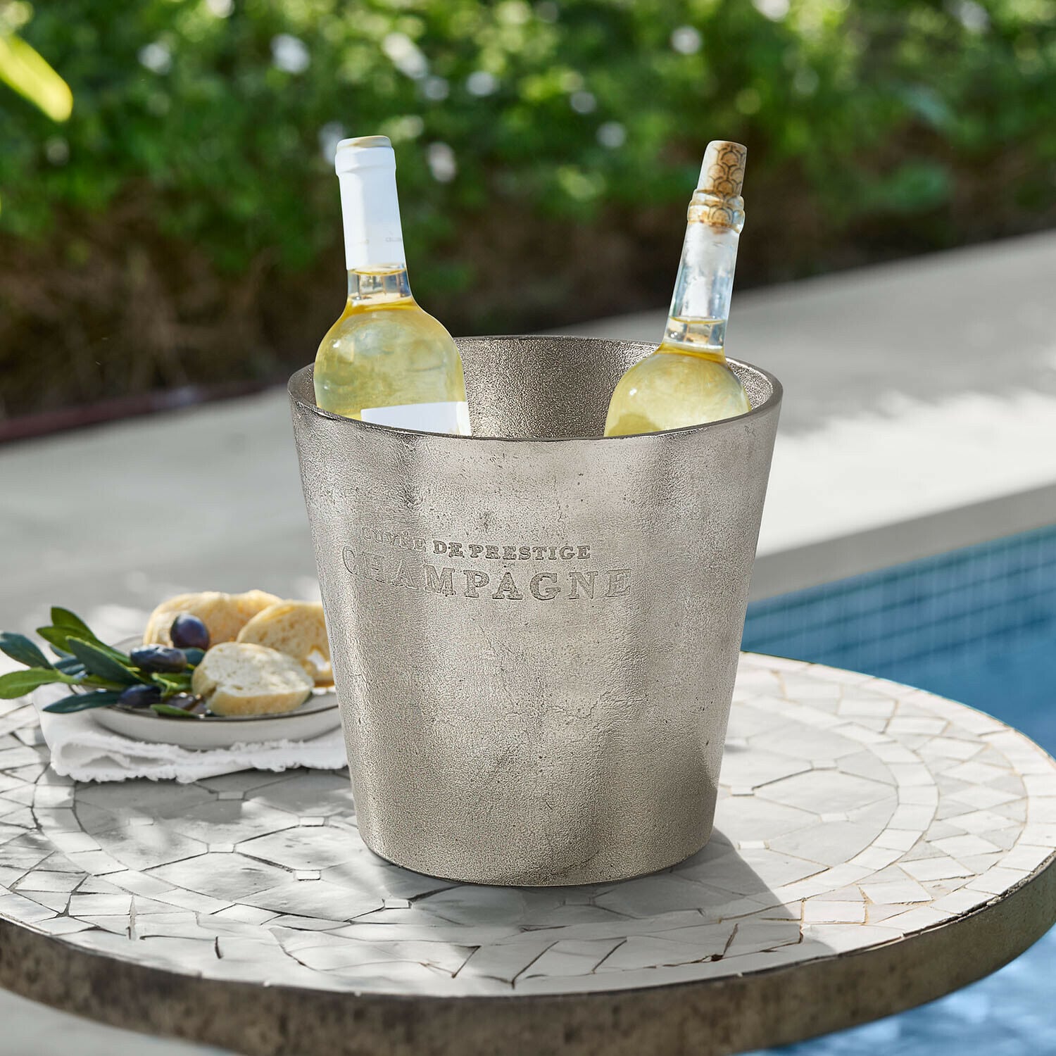 Metalowy srebrny kubeł na lód z napisem Champagne z dwoma butelkami białego wina na stole ogrodowym