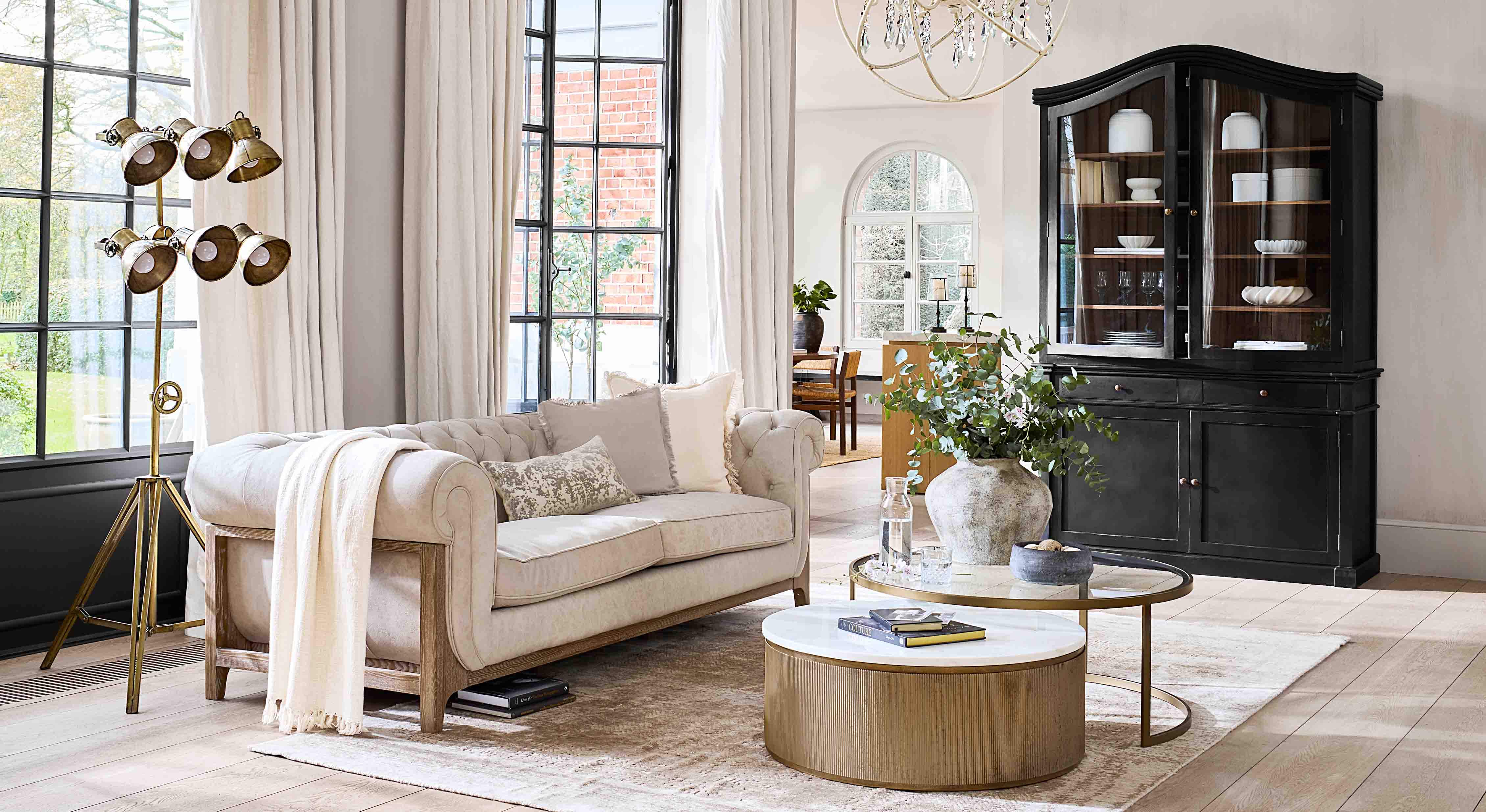 Beżowa sofa z drewnianą ramą w jasnym salonie z czarną witryną i złotą lampą stojącą