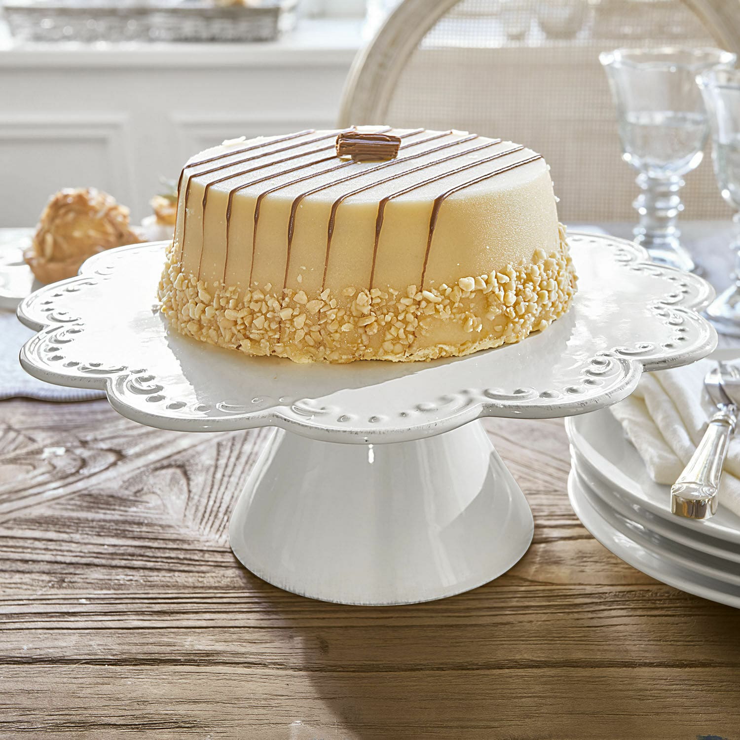 Patera na tort Fontienne Biały ceramiczny stojak na ciasto z ozdobnym brzegiem na drewnianym stole w jadalni