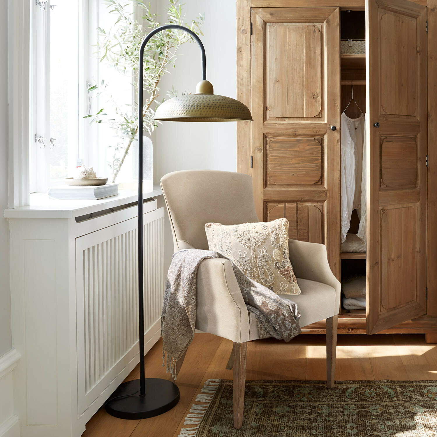 Beżowy fotel z drewnianymi nogami, ozdobna poduszka i pled, obok metalowa lampa podłogowa z mosiężnym kloszem