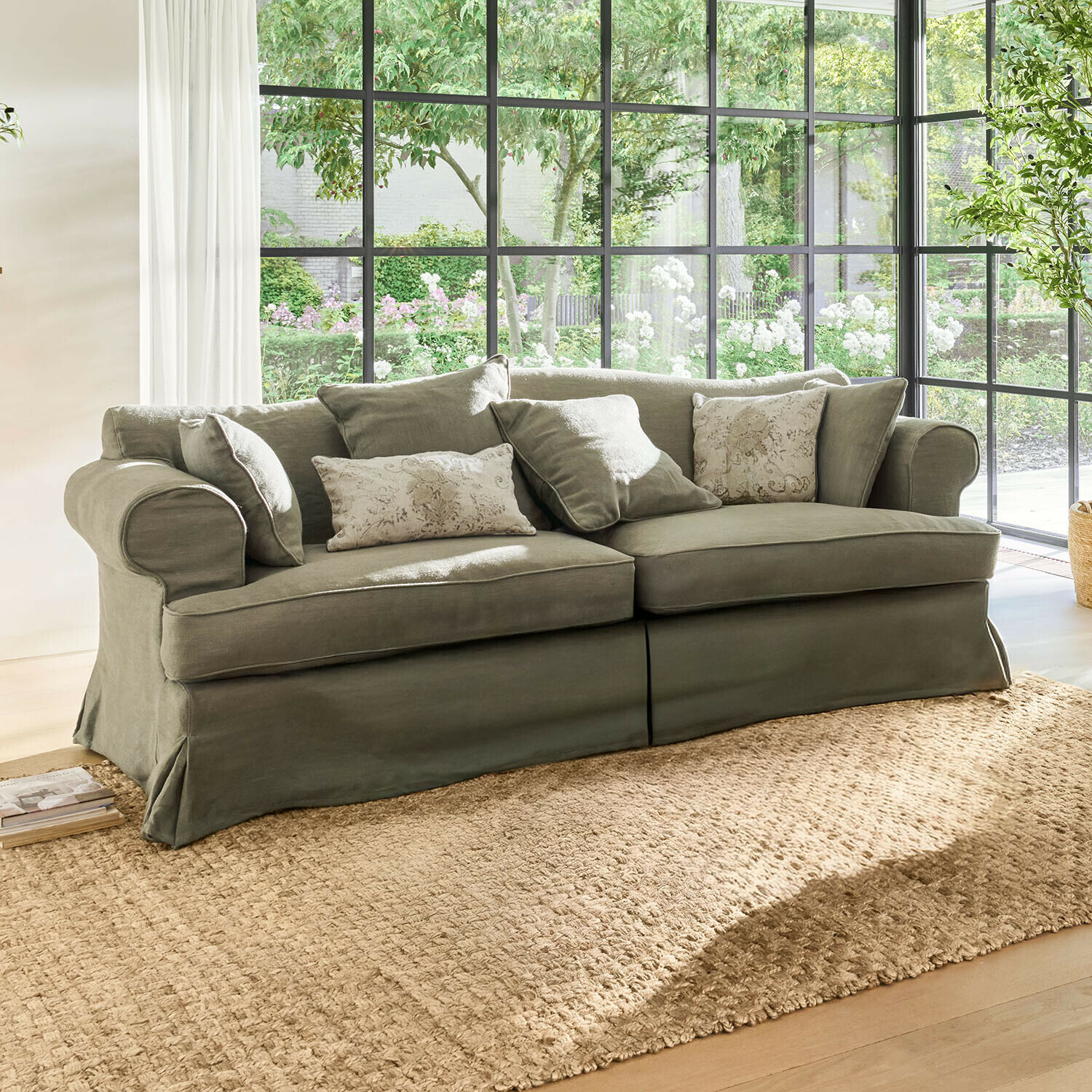 Sofa Dawnhurst Szara sofa z tkaniny z poduszkami dekoracyjnymi na tle dużych okien i naturalnego światła