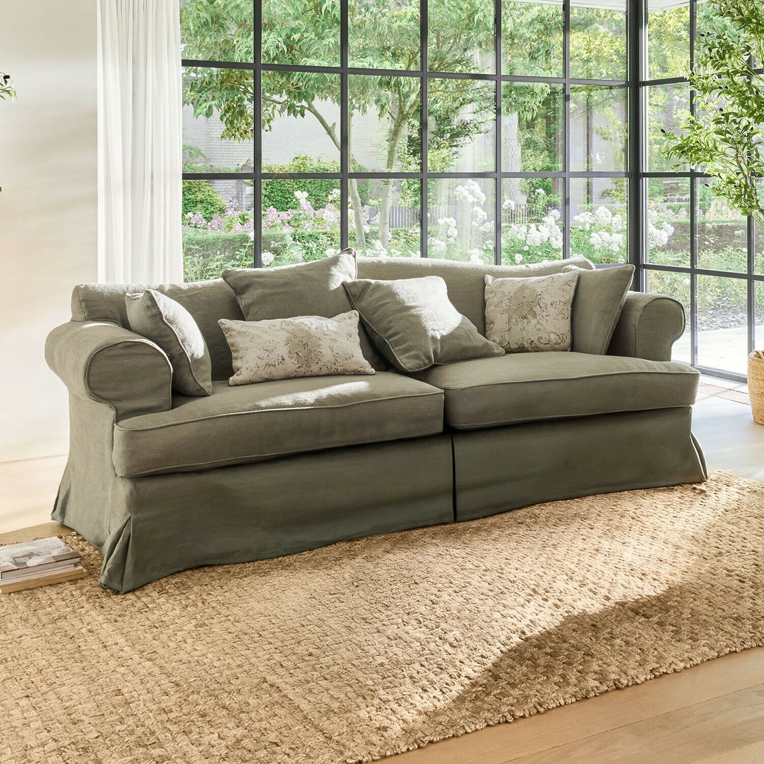 Sofa Dawnhurst Szara sofa z tkaniny z poduszkami dekoracyjnymi na tle dużych okien i naturalnego światła