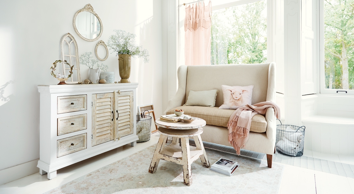 Przytulny salon w stylu shabby chic z białą drewnianą komodą i beżową sofą z poduszkami i różowym kocem