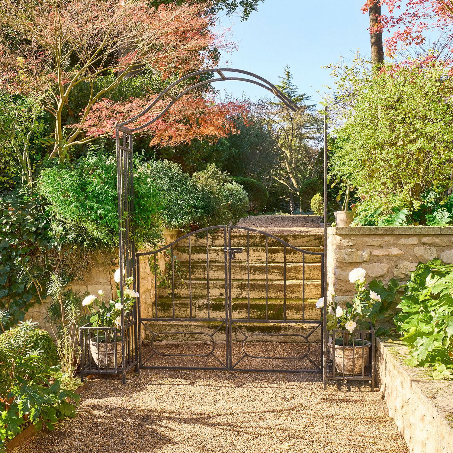 Ozdobna czarna metalowa brama ogrodowa z łukiem i donicami z białymi kwiatami na żwirowej ścieżce