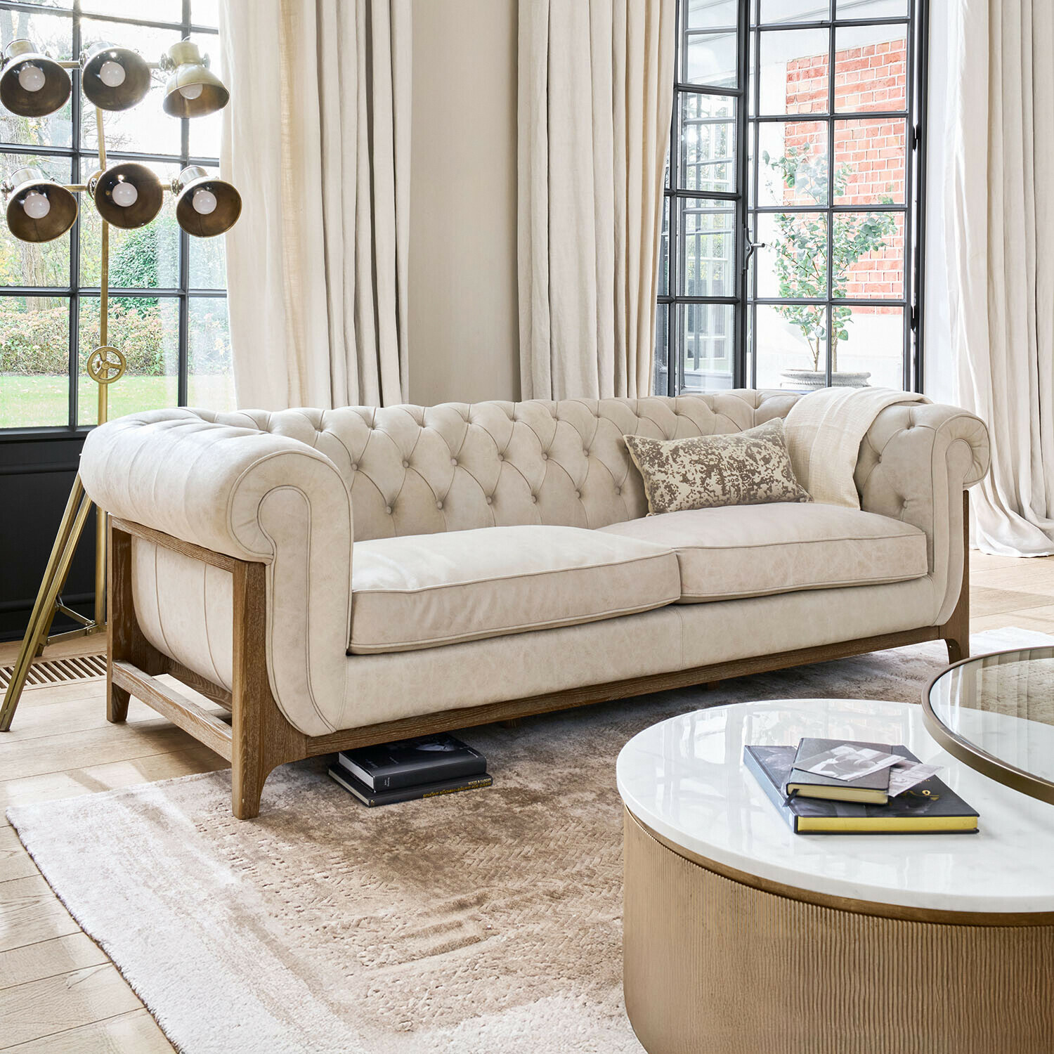 Beżowa pikowana sofa z drewnianą ramą w jasnym salonie z dużymi oknami i lampą stojącą