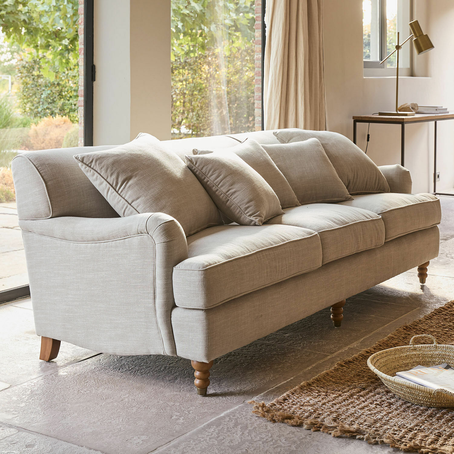 Sofa Isa Beżowa sofa tapicerowana z poduszkami i drewnianymi nogami w jasnym salonie z dywanem