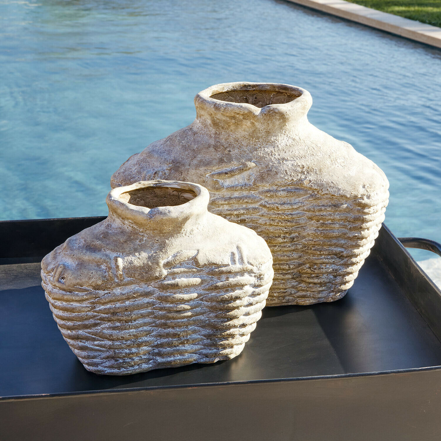 Dwie dekoracyjne wazony z kamienia w beżowym kolorze na czarnej tacy przy basenie