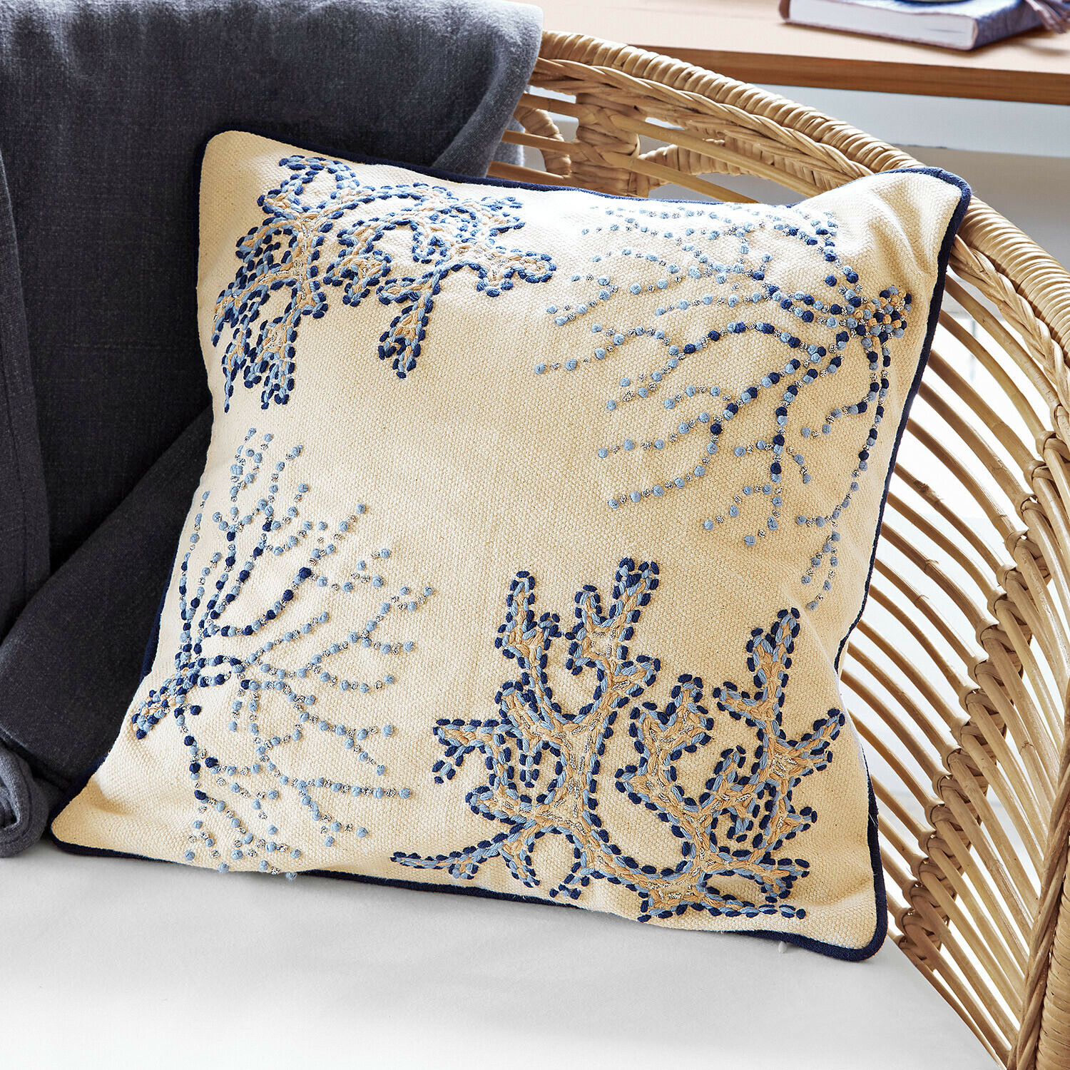 Beżowa poduszka dekoracyjna z niebieskim haftem i granatową lamówką na wiklinowym fotelu
