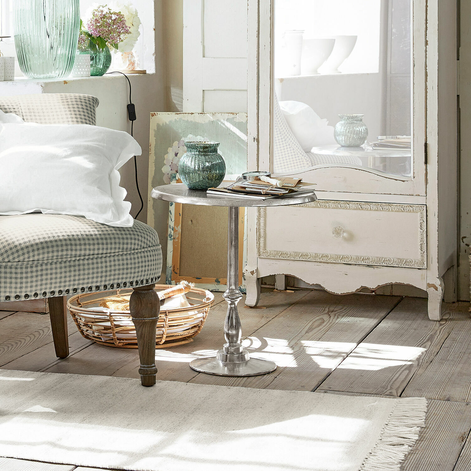 Metalowy okrągły stolik boczny w srebrnym kolorze w jasnym salonie z wiklinowym koszem i beżowym fotelem