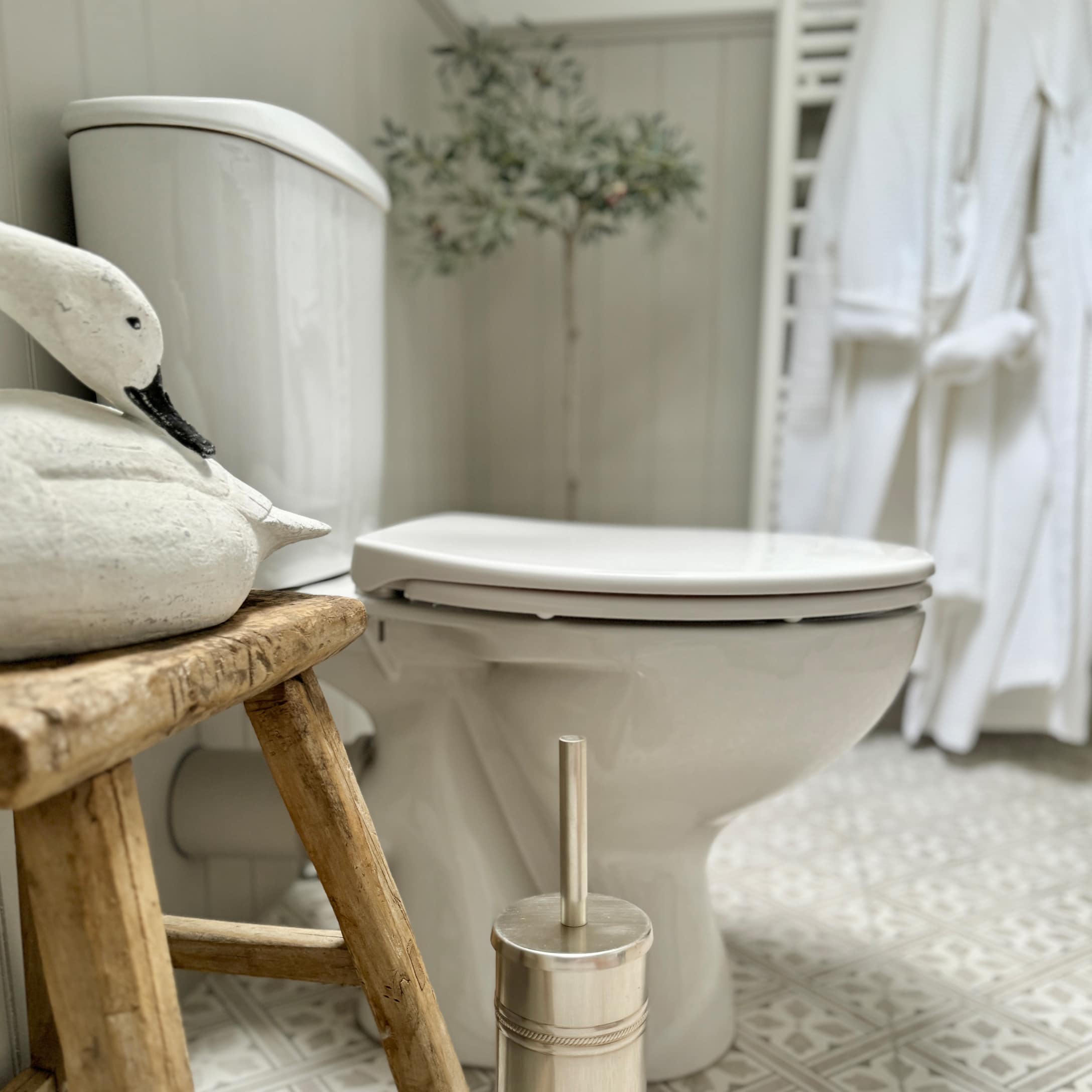 Biała ceramiczna toaleta z drewnianym stołkiem i metalową szczotką łazienkową na wzorzystej podłodze