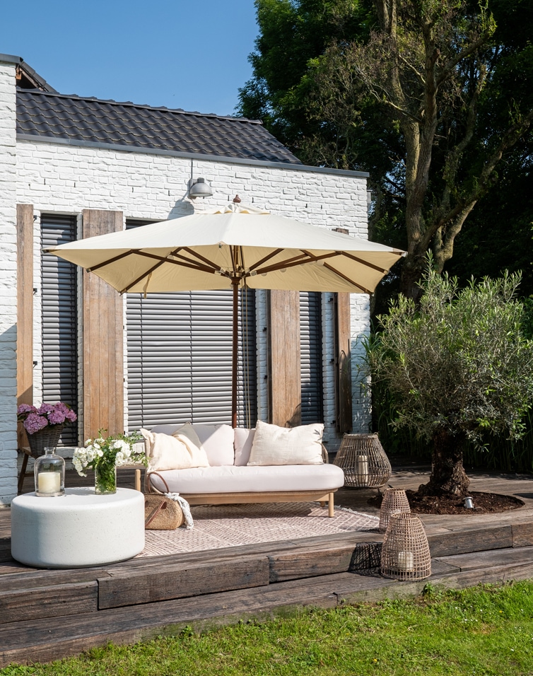 Taras z białą cegłą, beżowa sofa z poduszkami i kremowy parasol ogrodowy na drewnianym tarasie