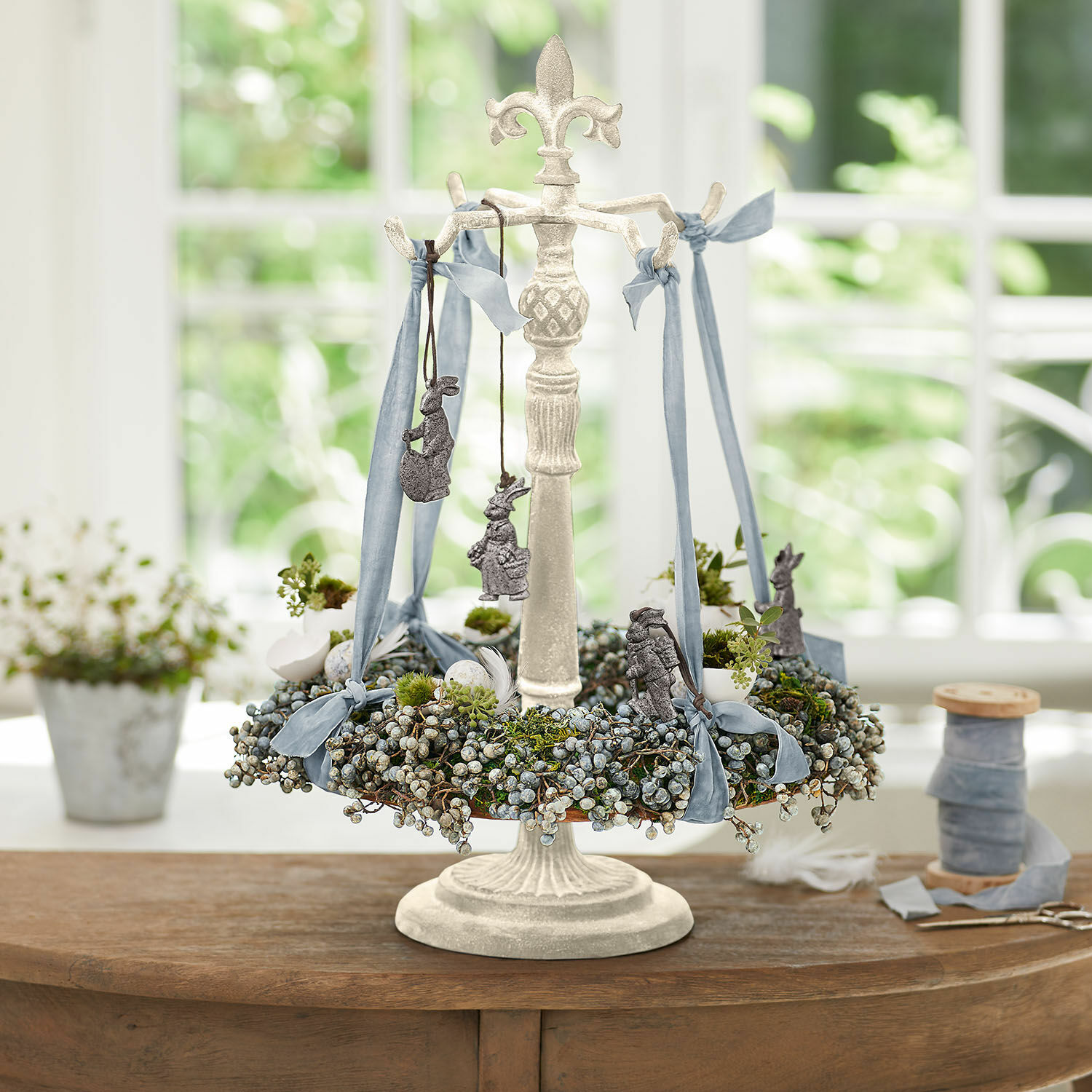 Stojak na wieniec Antwan Dekoracyjny stojak z białego metalu z zielonym wieńcem i niebieskimi wstążkami na stół wiosenny