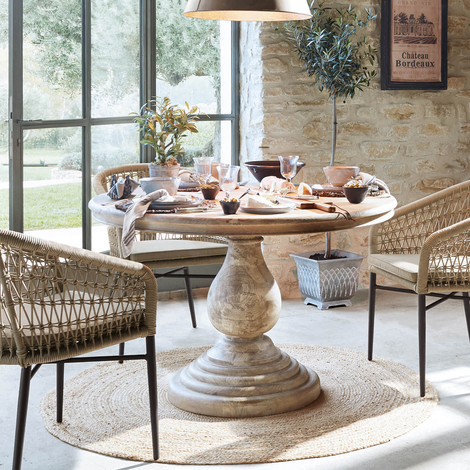 Okrągły drewniany stół z masywną podstawą na jasnym dywanie, otoczony wiklinowymi krzesłami w jadalni