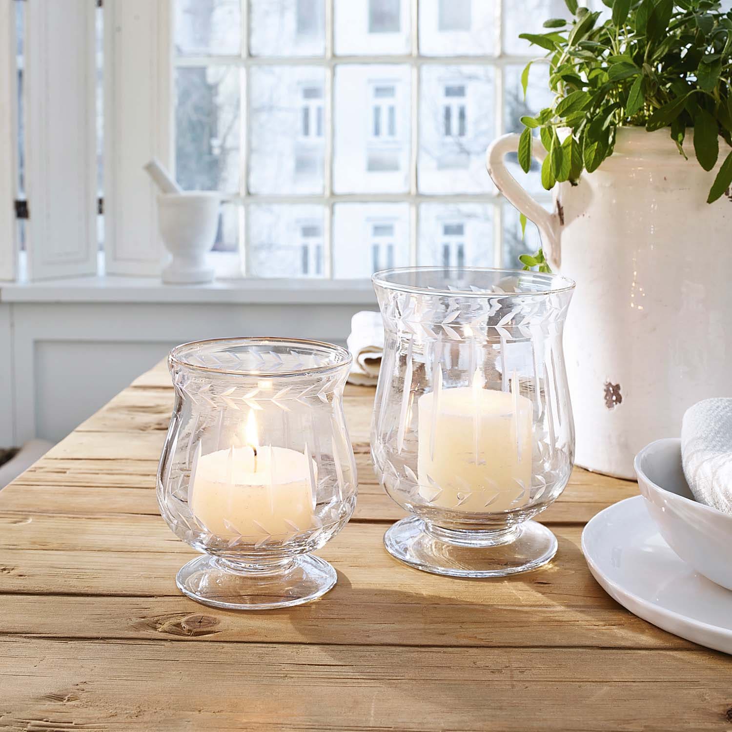 Dwa szklane świeczniki z białymi świecami na drewnianym stole w jasnym wnętrzu