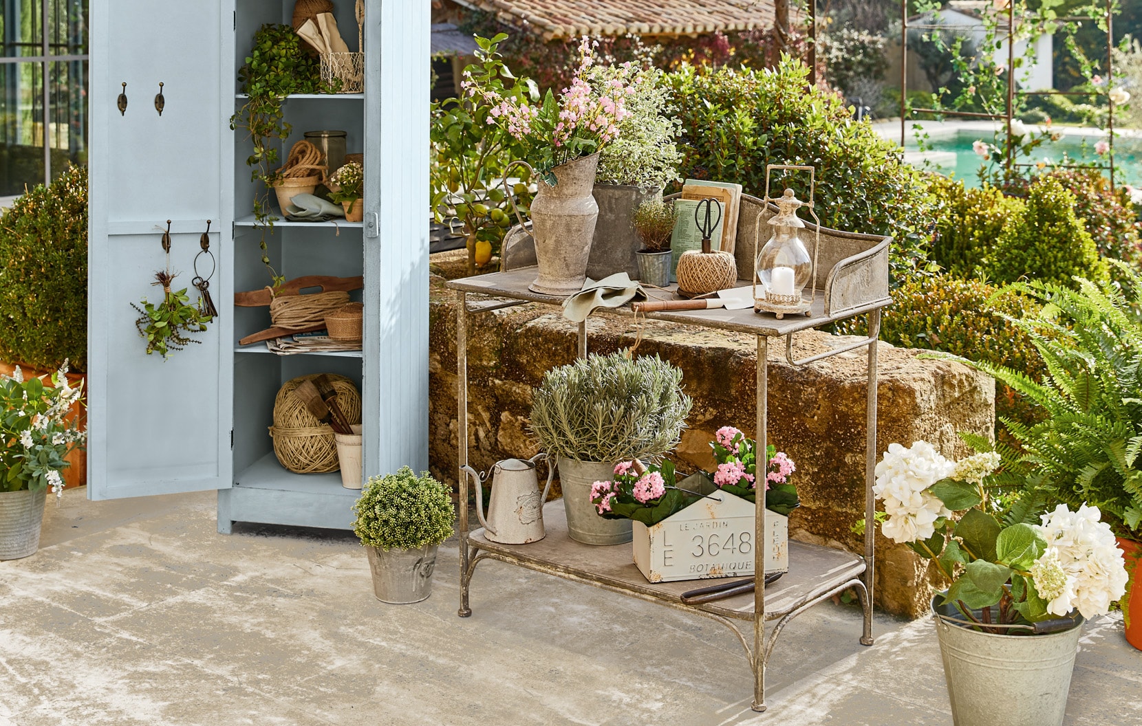 Metalowy wózek ogrodowy z dekoracjami i kwiatami w stylu shabby chic na patio z szafką.