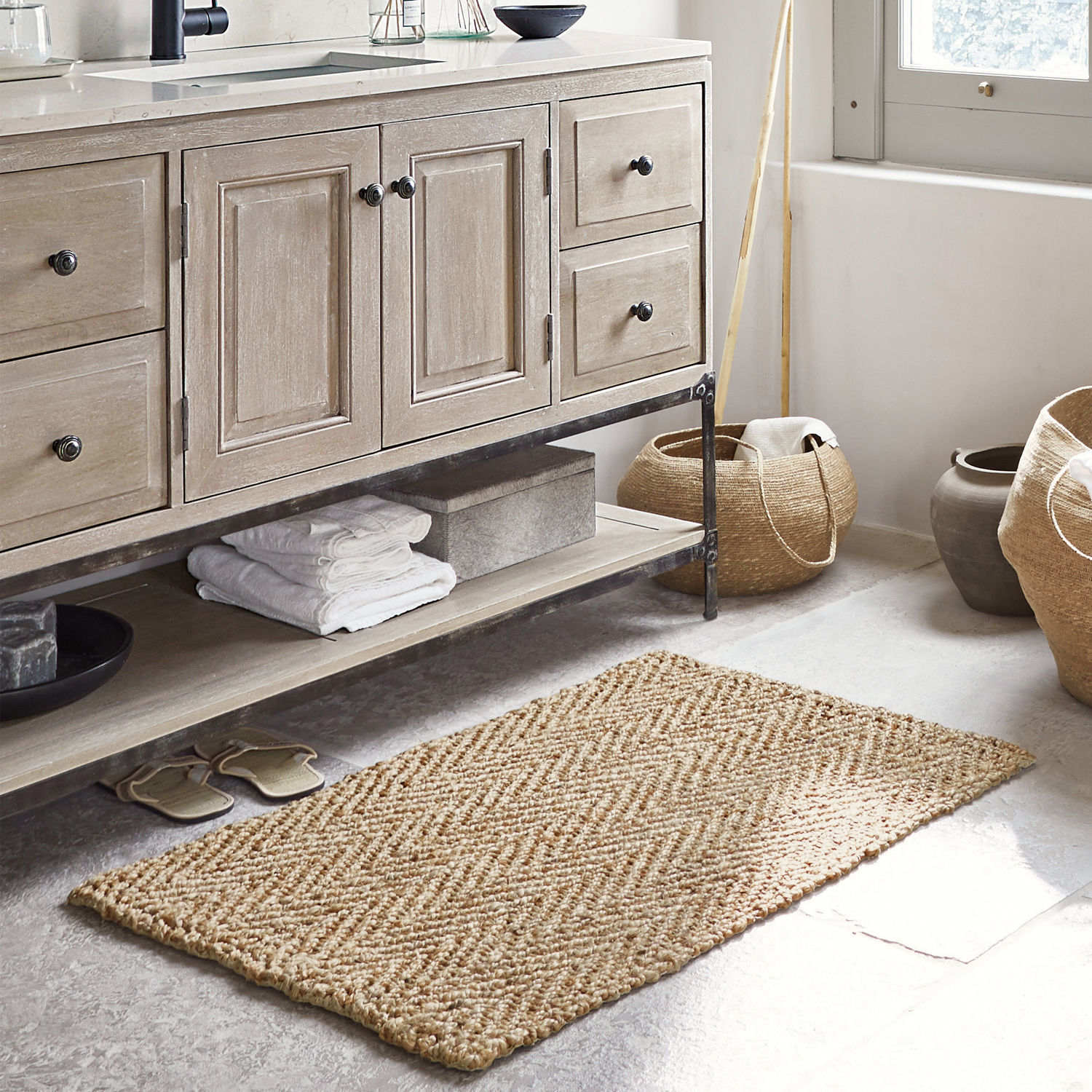 Dywanik łazienkowy Daoukro Beżowy dywanik z juty o splocie w jodełkę na podłodze łazienki z drewnianą szafką i koszami