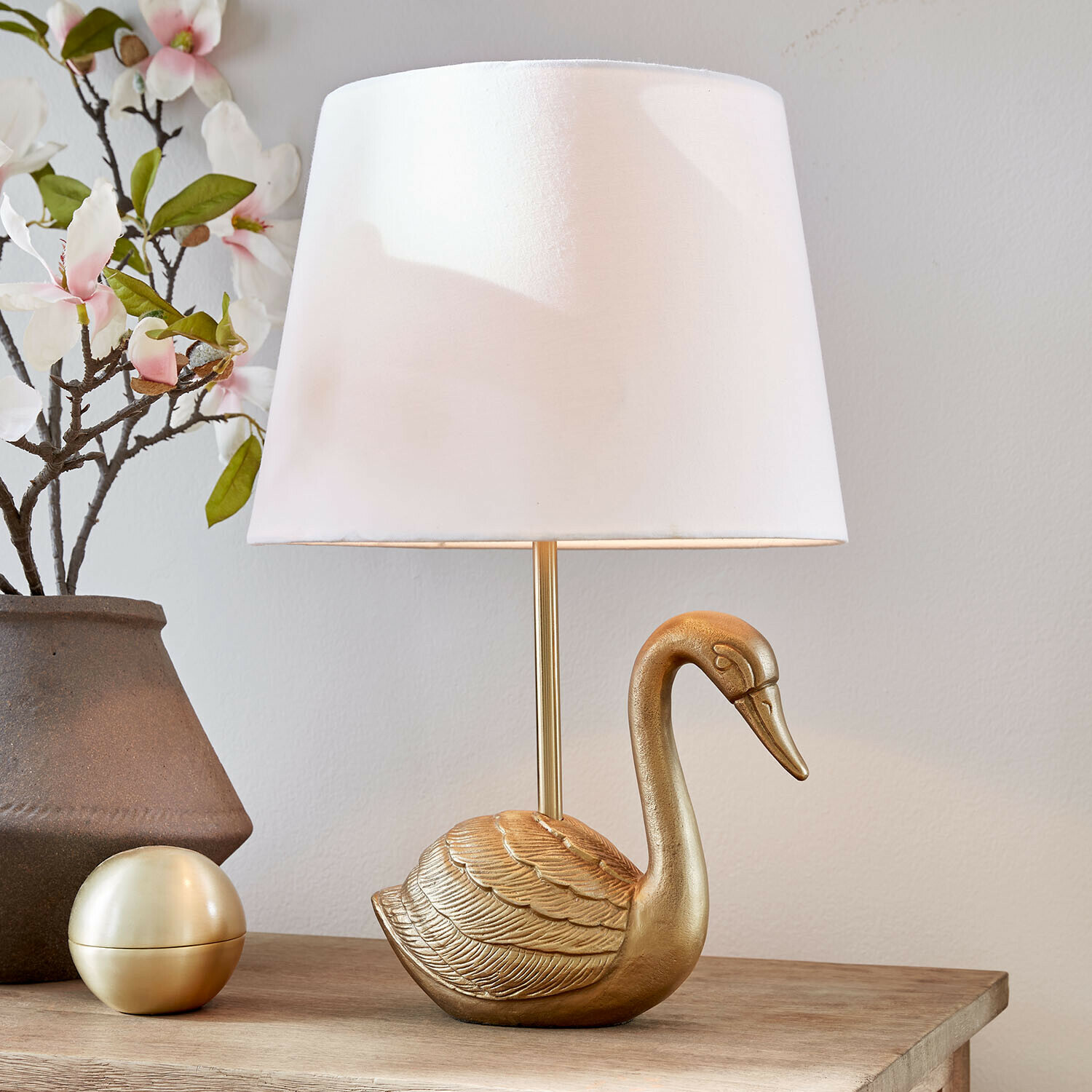 Lampa stołowa Élégance Dorée Złota lampka stołowa w kształcie łabędzia z białym abażurem na drewnianym stole z dekoracją kwiatową