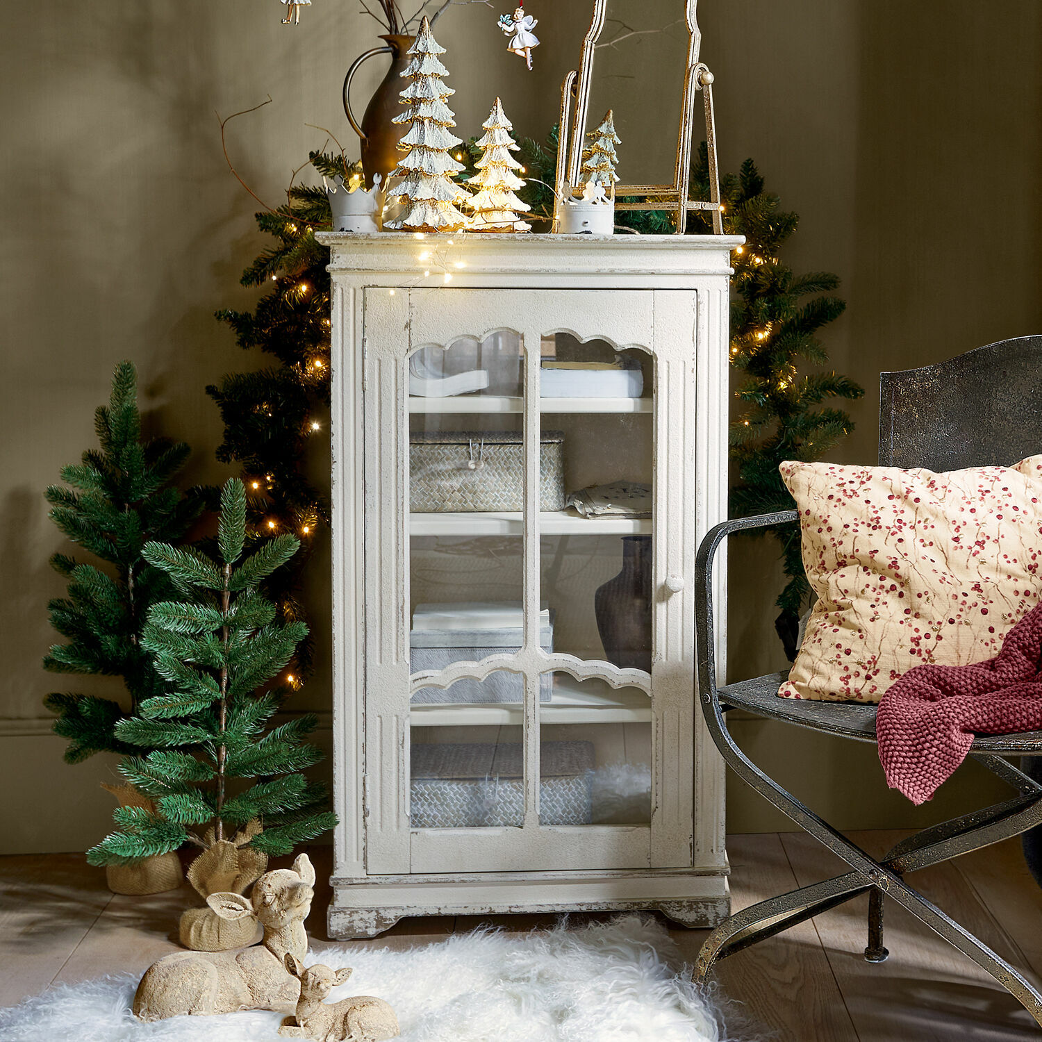 Biała drewniana szafka z przeszklonymi drzwiami w stylu vintage, dekoracje świąteczne i zielone drzewka