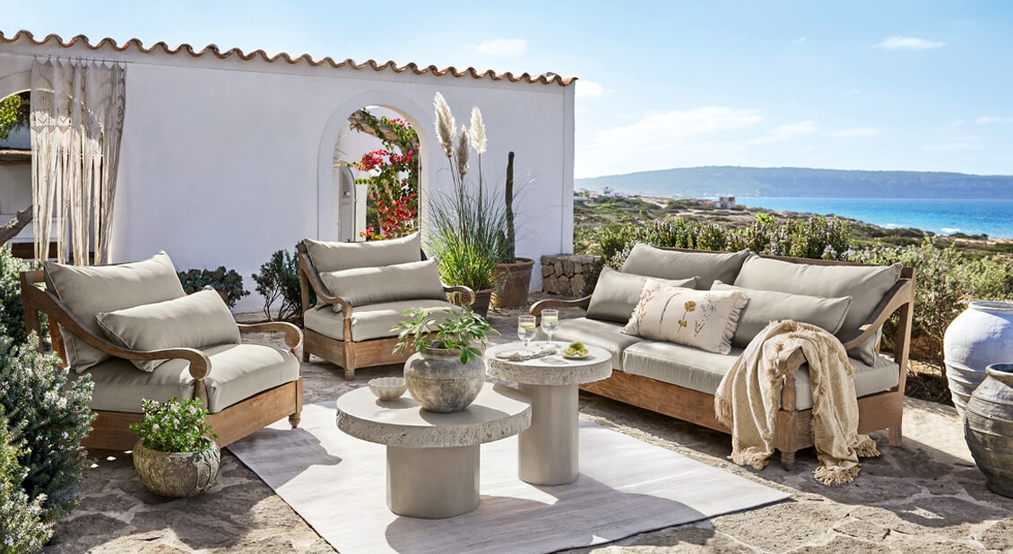 Zestaw mebli ogrodowych z drewnianą ramą i beżowymi poduszkami na kamiennym tarasie z widokiem na morze