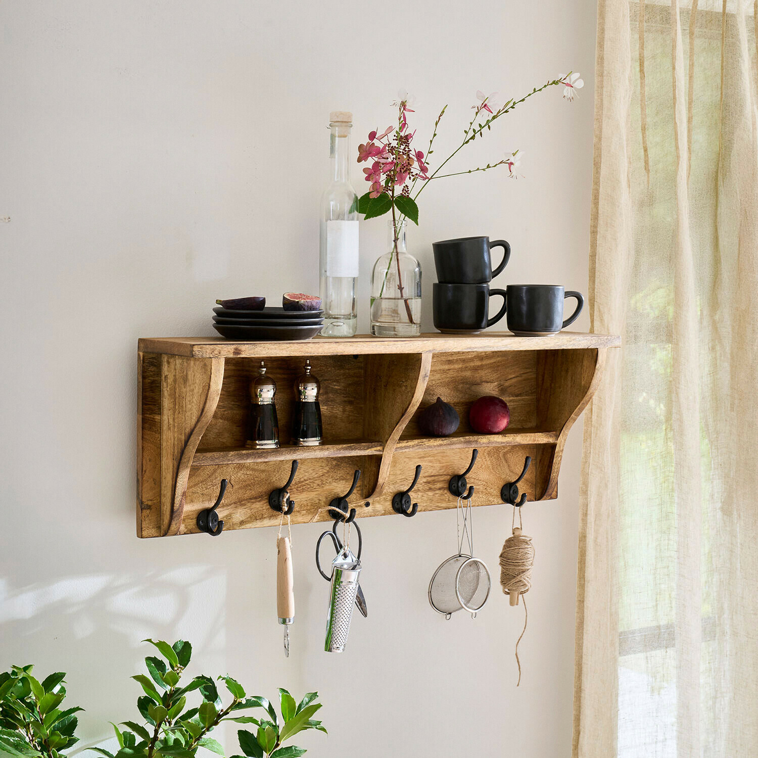 Drewniana półka ścienna z haczykami i czarnymi kubkami, talerzami oraz dekoracją kwiatową w jasnym wnętrzu