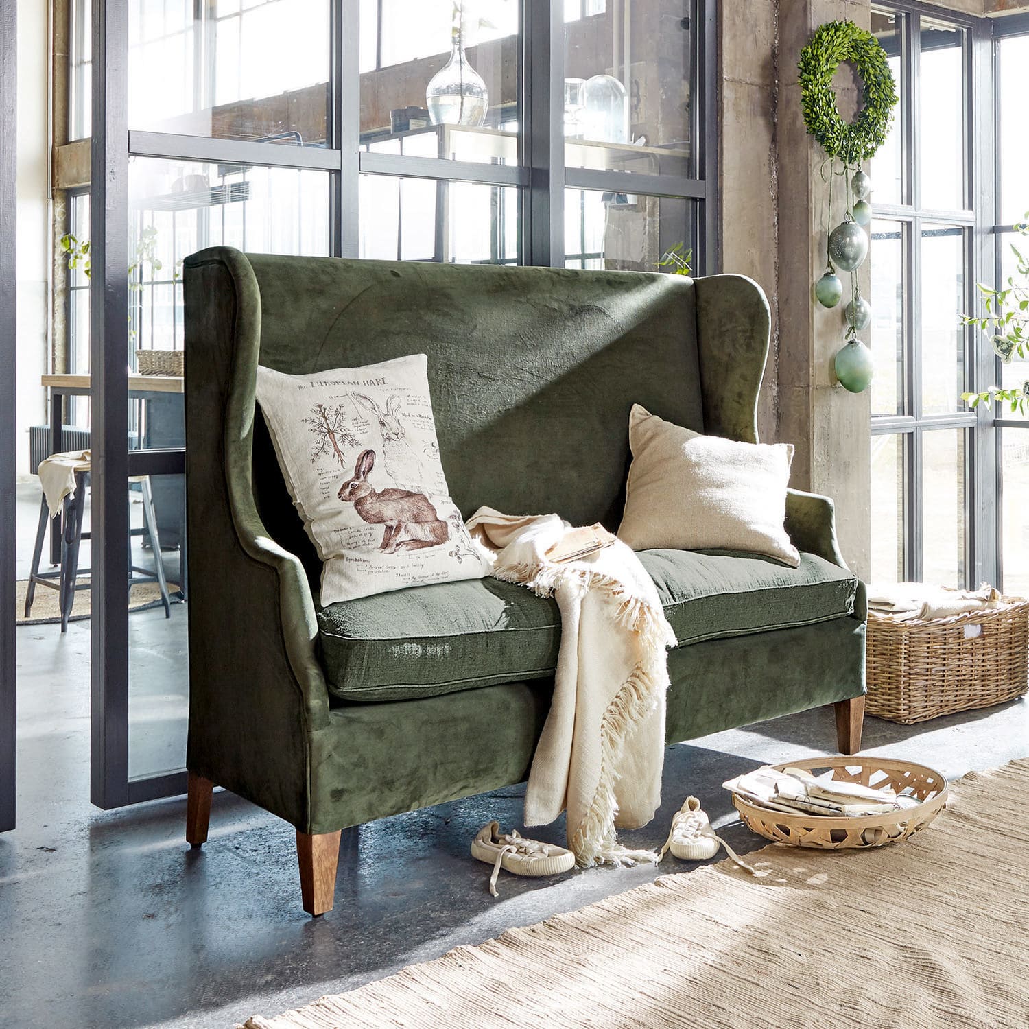 Sofa Ombra Zielona aksamitna sofa z poduszkami i kremowym kocem w jasnym, nowoczesnym wnętrzu z dużymi oknami