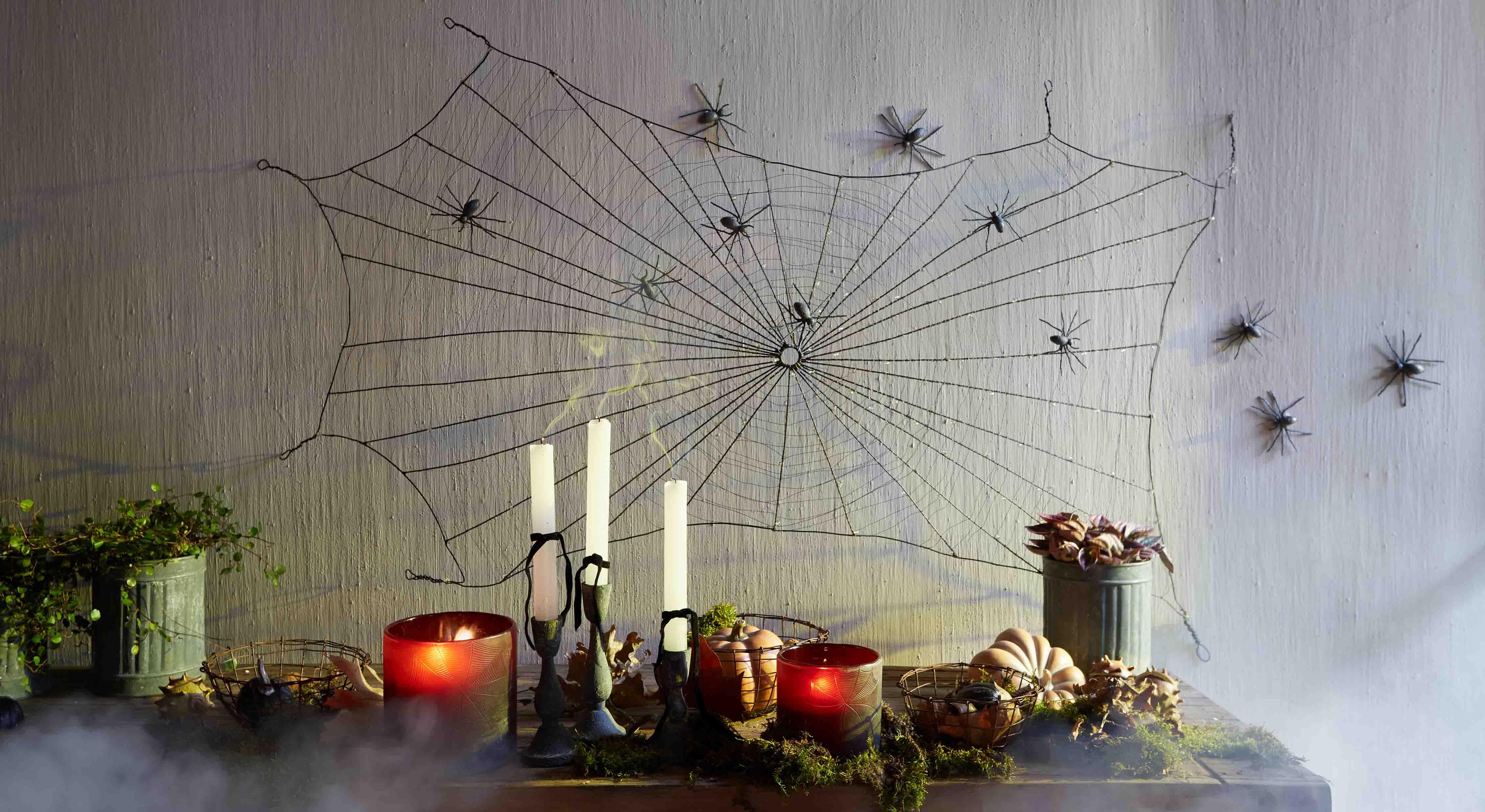 Dekoracja Halloween z czarną pajęczyną i plastikowymi pająkami na ścianie oraz świecami i dyniami na stole