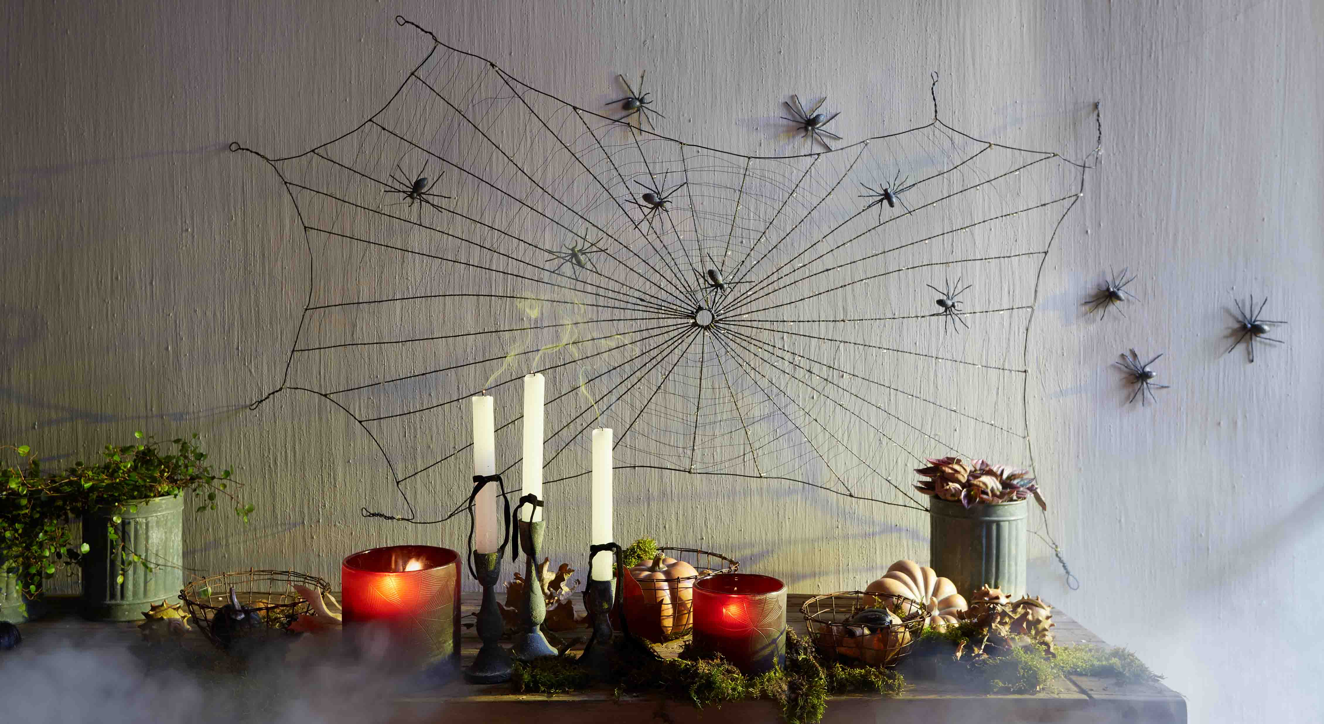 Dekoracja Halloween z czarną pajęczyną i plastikowymi pająkami na ścianie oraz świecami i dyniami na stole