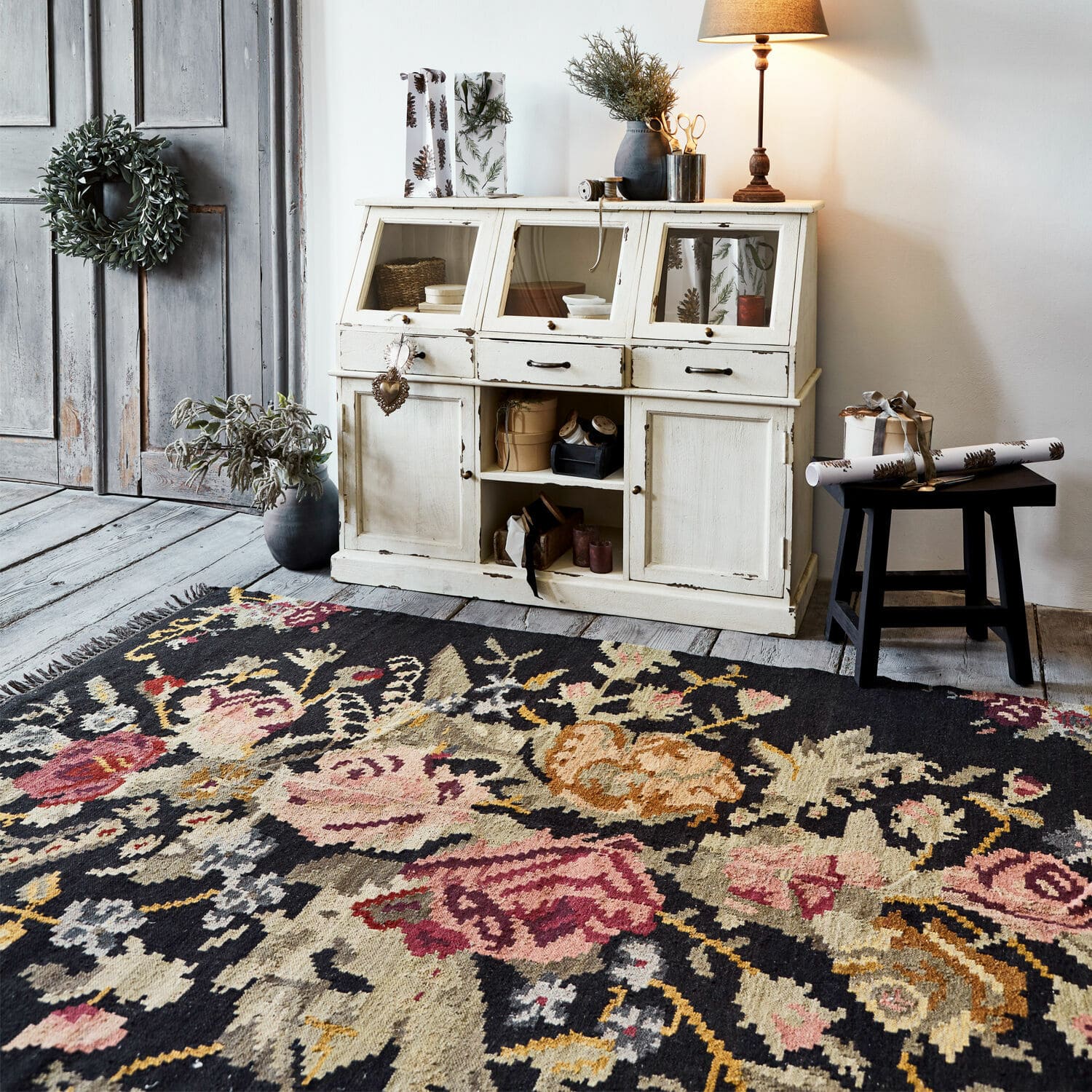 Dywan z kwiatowym wzorem w ciemnych kolorach na tle białej drewnianej komody i lampy stołowej