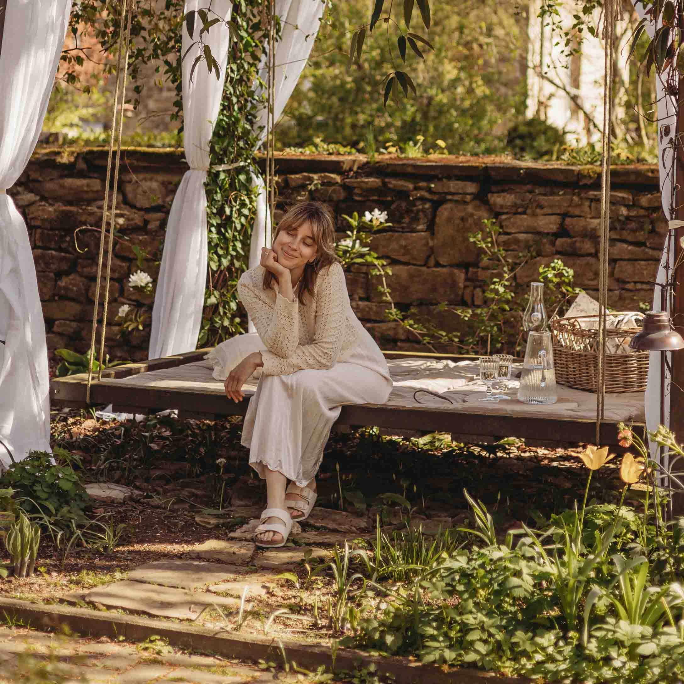 Kobieta w beżowym swetrze siedzi na huśtawce ogrodowej z drewnianą ramą i białymi zasłonami