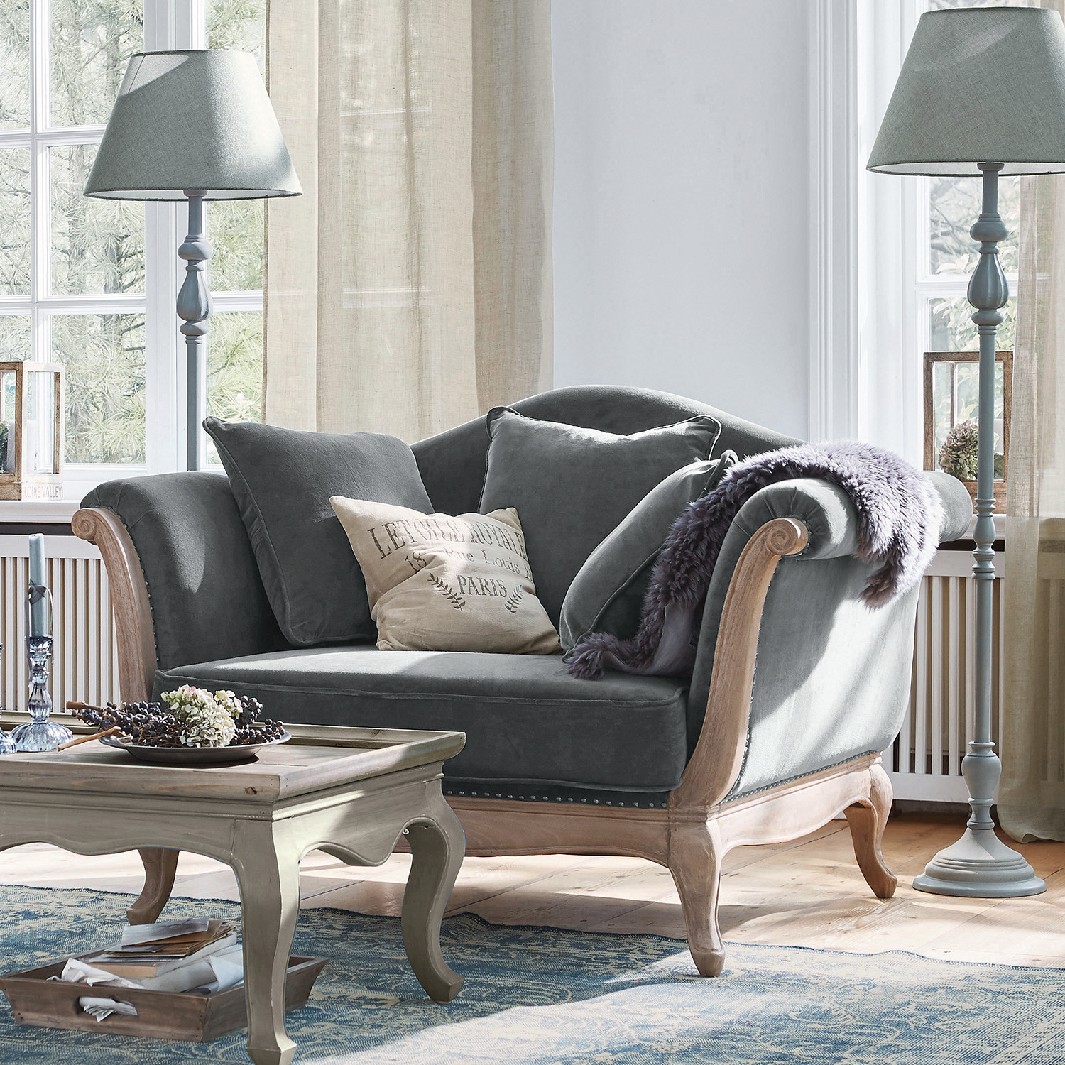 Fotel Sumerset Szara aksamitna sofa z drewnianą ramą i poduszkami w jasnym salonie z lampami stojącymi