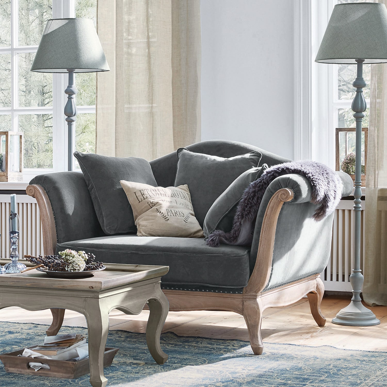 Fotel Sumerset Szara aksamitna sofa z drewnianą ramą i poduszkami w jasnym salonie z lampami stojącymi