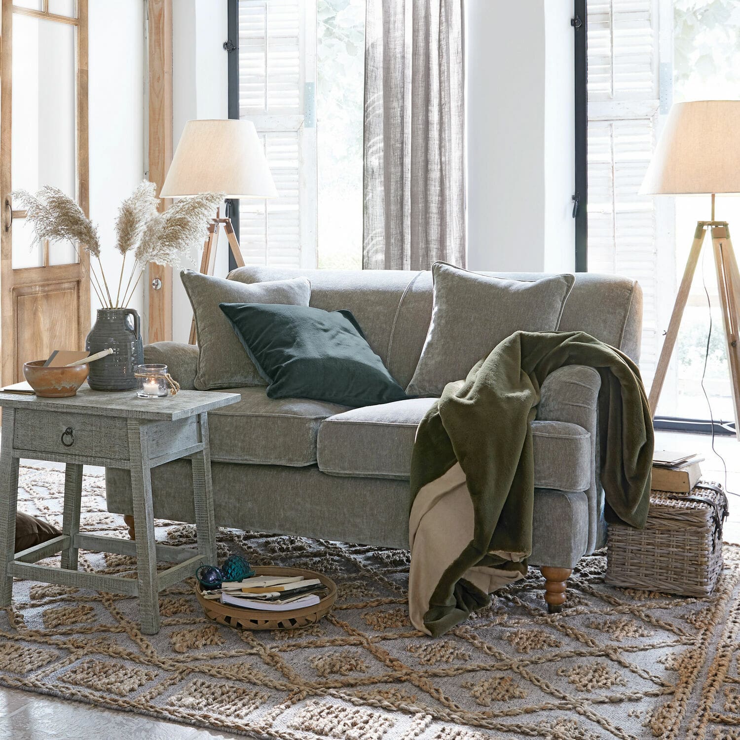 Szara sofa z miękkimi poduszkami i zielonym kocem w jasnym salonie z drewnianymi lampami i dywanem