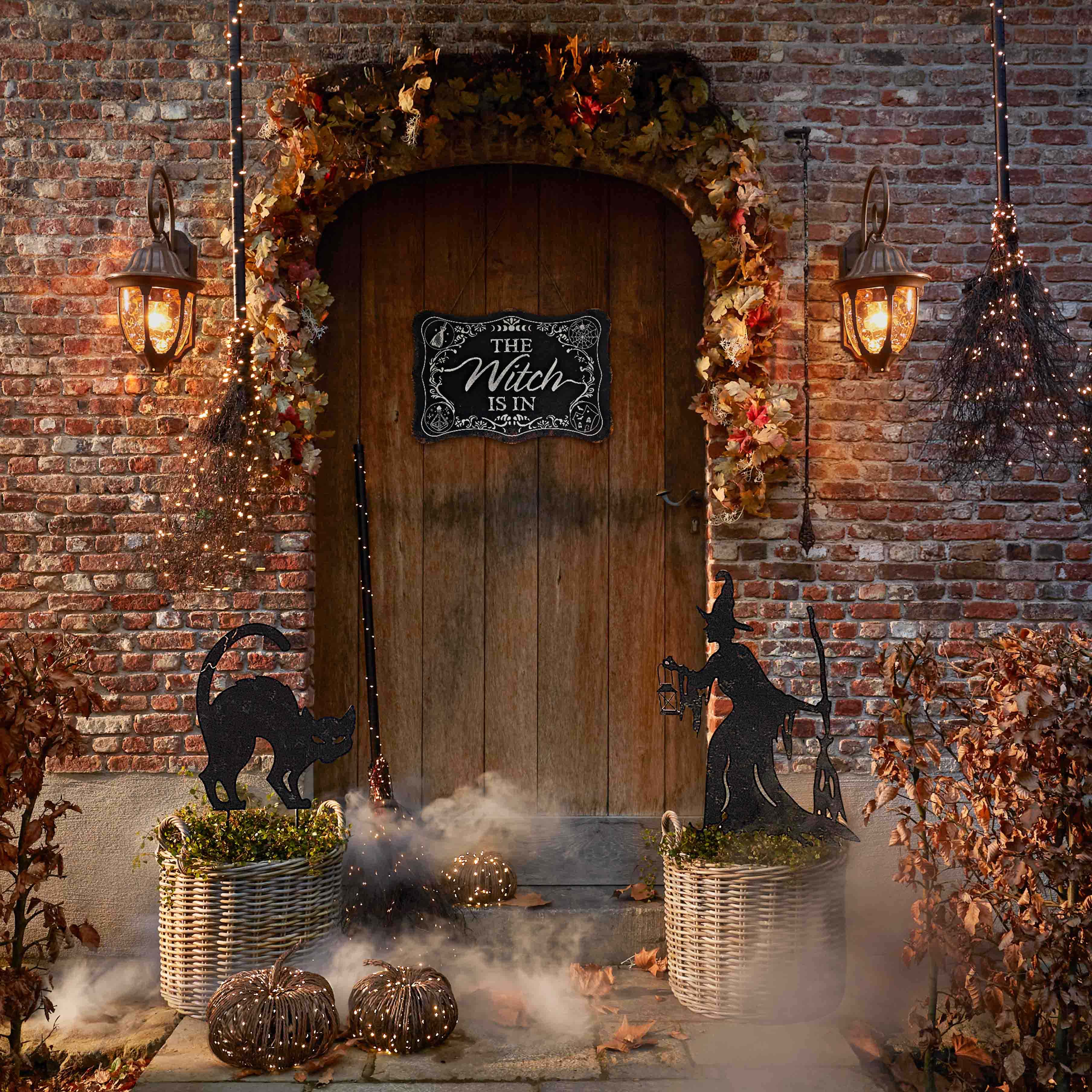 Dekoracja halloweenowa z czarną kotką, czarownicą i lampionami przy drewnianych drzwiach z cegły
