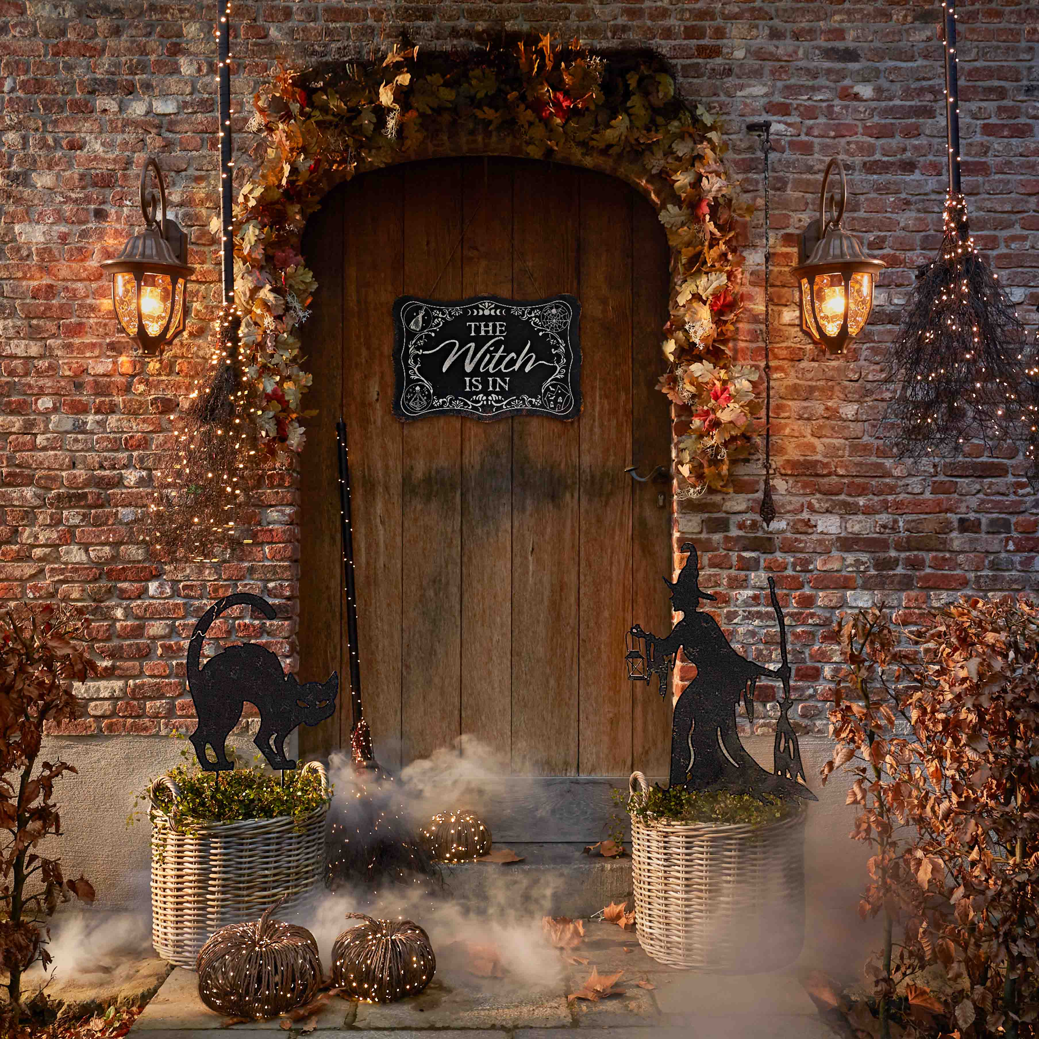 Dekoracja halloweenowa z czarną kotką, czarownicą i lampionami przy drewnianych drzwiach z cegły