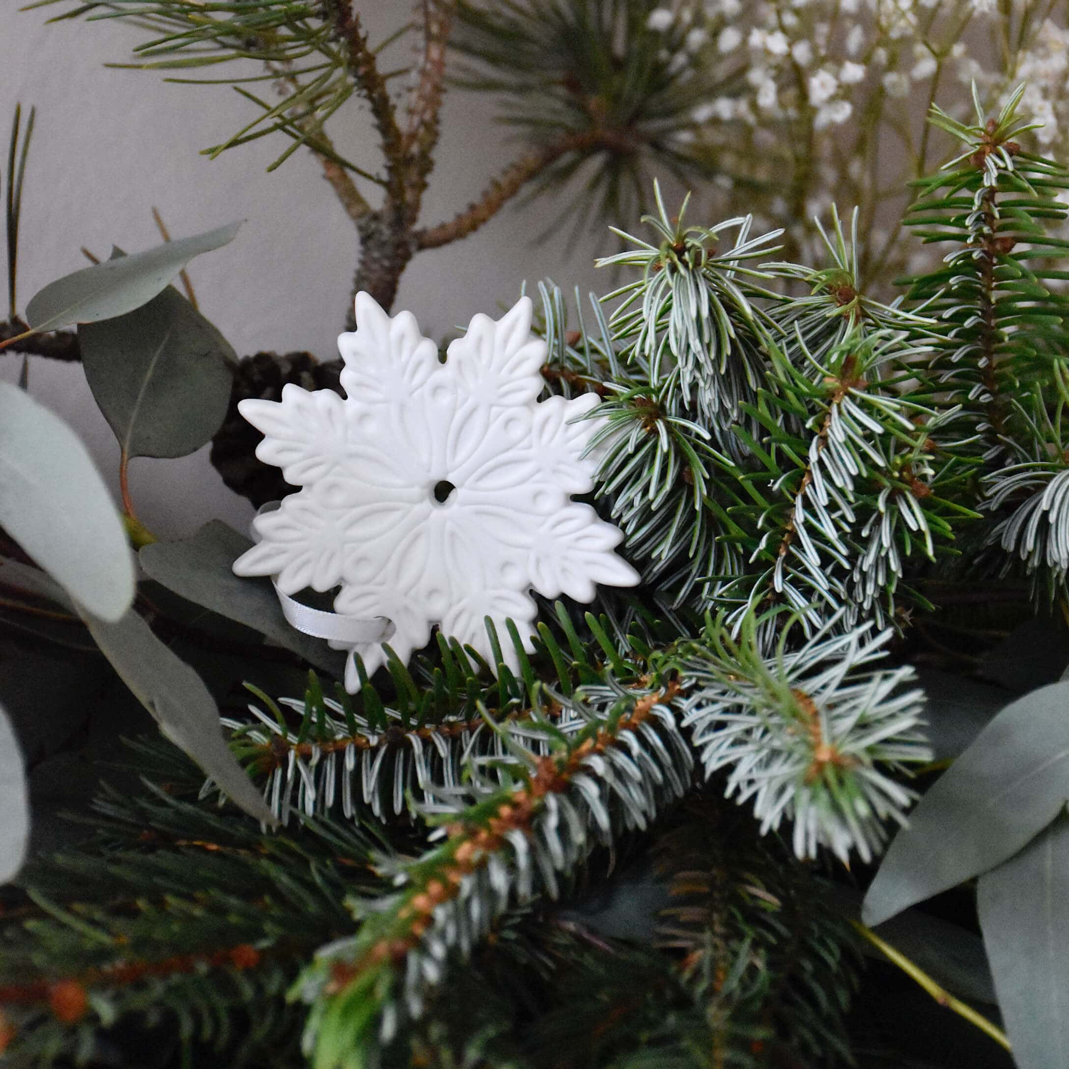 Biała ceramiczna ozdoba świąteczna w kształcie płatka śniegu na zielonych gałązkach świerku
