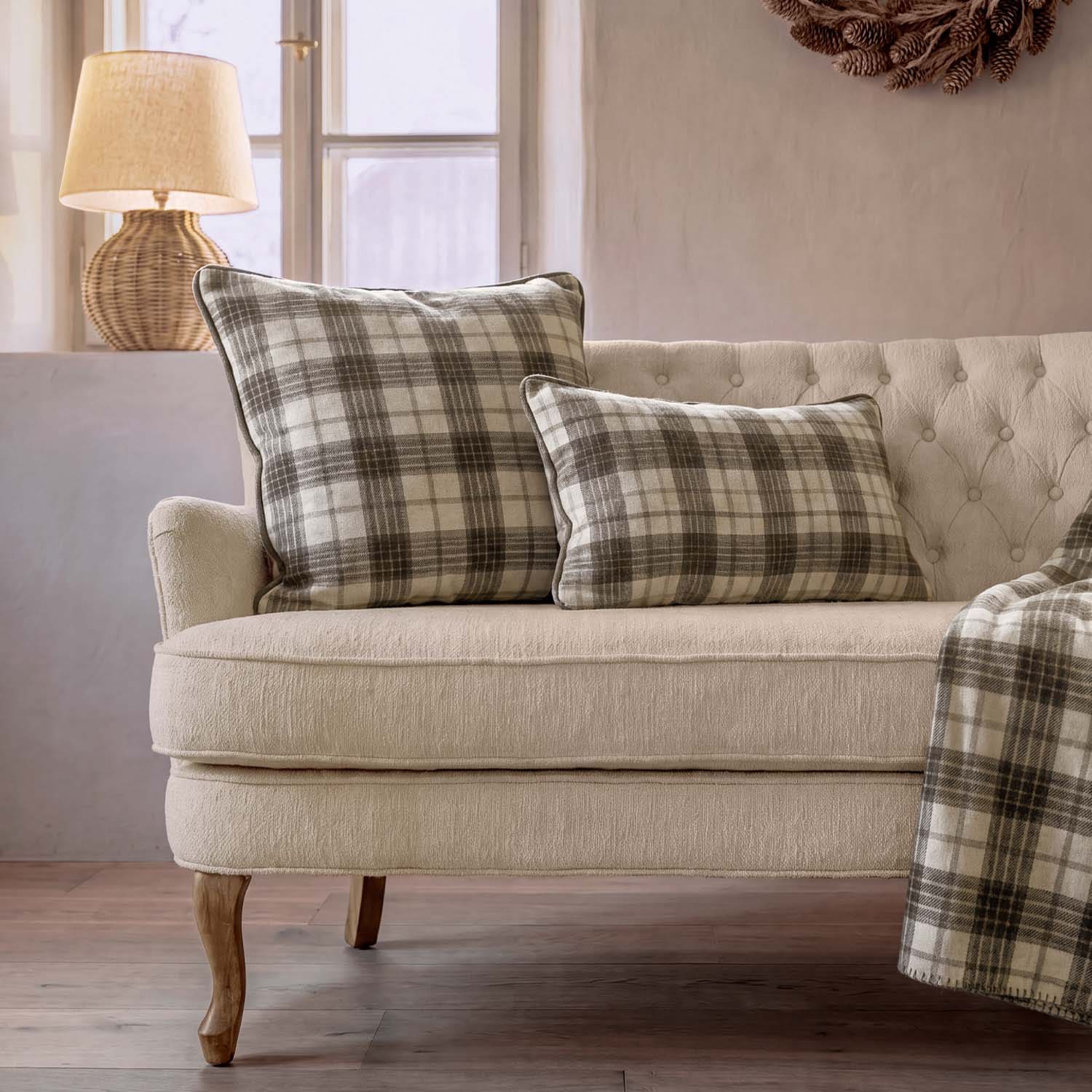 Beżowa sofa z dwoma poduszkami w kratę w odcieniach szarości i kremu oraz lampą na tle okna