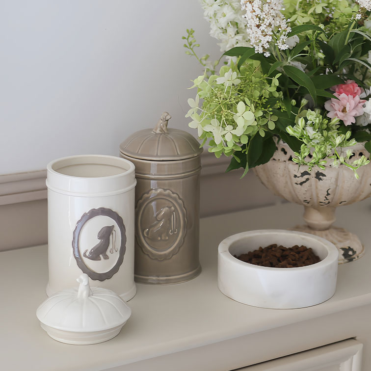 Ceramiczne pojemniki na karmę dla psa w kremowym i beżowym kolorze obok białej miski z karmą i kwiatów