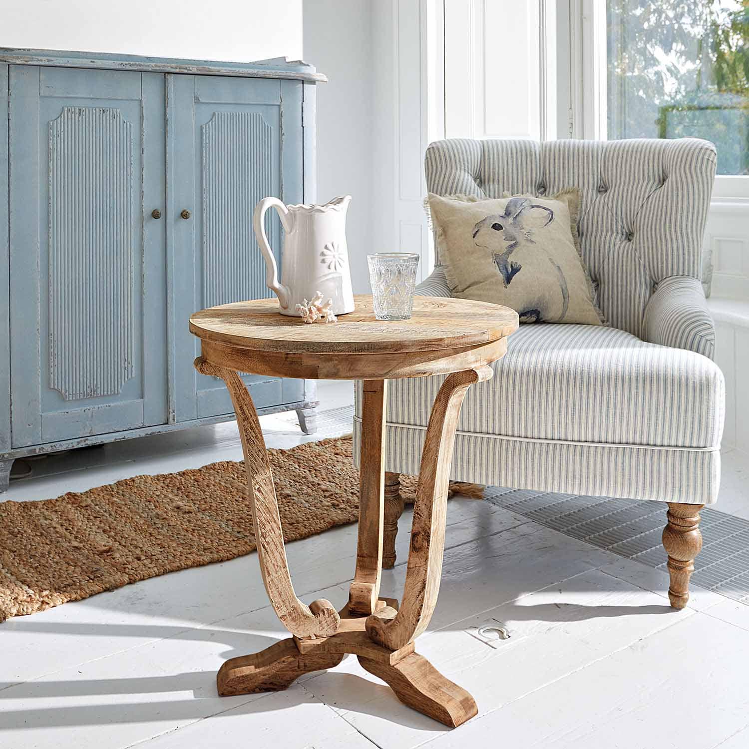 Okrągły stolik z drewna mango w naturalnym kolorze w jasnym salonie z fotelami i niebieską komodą