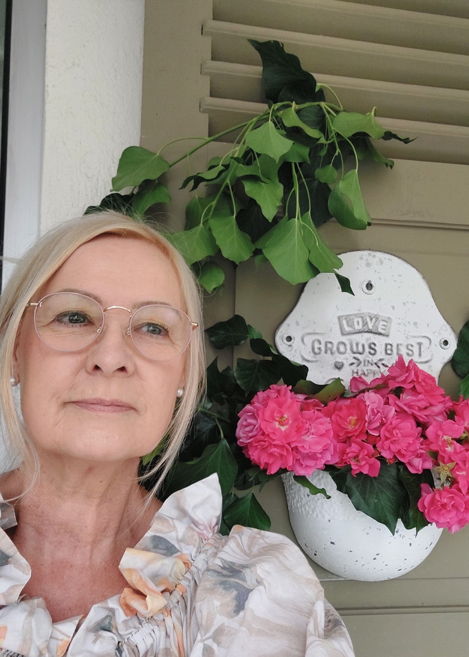 Biała ceramiczna doniczka z różowymi kwiatami i zielonymi liśćmi na tle szarych żaluzji
