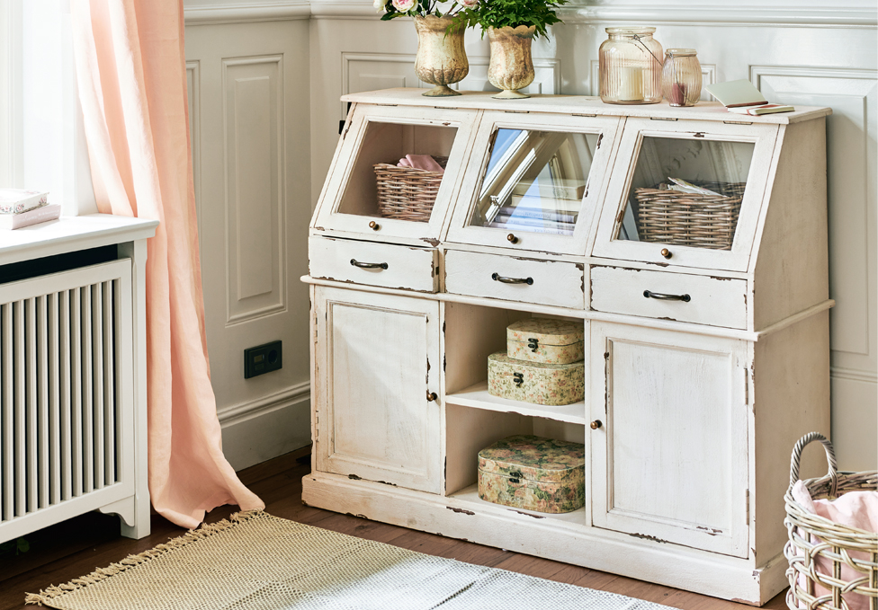 Biały drewniany kredens w stylu shabby chic z przeszklonymi szufladami i dekoracjami w pokoju.