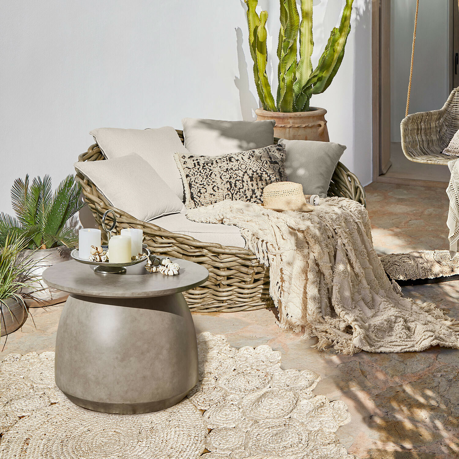 Okrągła sofa z rattanu z kremowymi poduszkami i pledem na tarasie z betonowym stolikiem