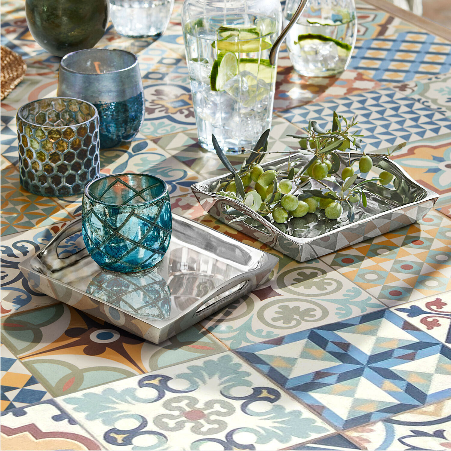 Dwustronne metalowe tace srebrne na kolorowym stole z mozaiką, dekoracyjne szklane świeczniki i zielone oliwki