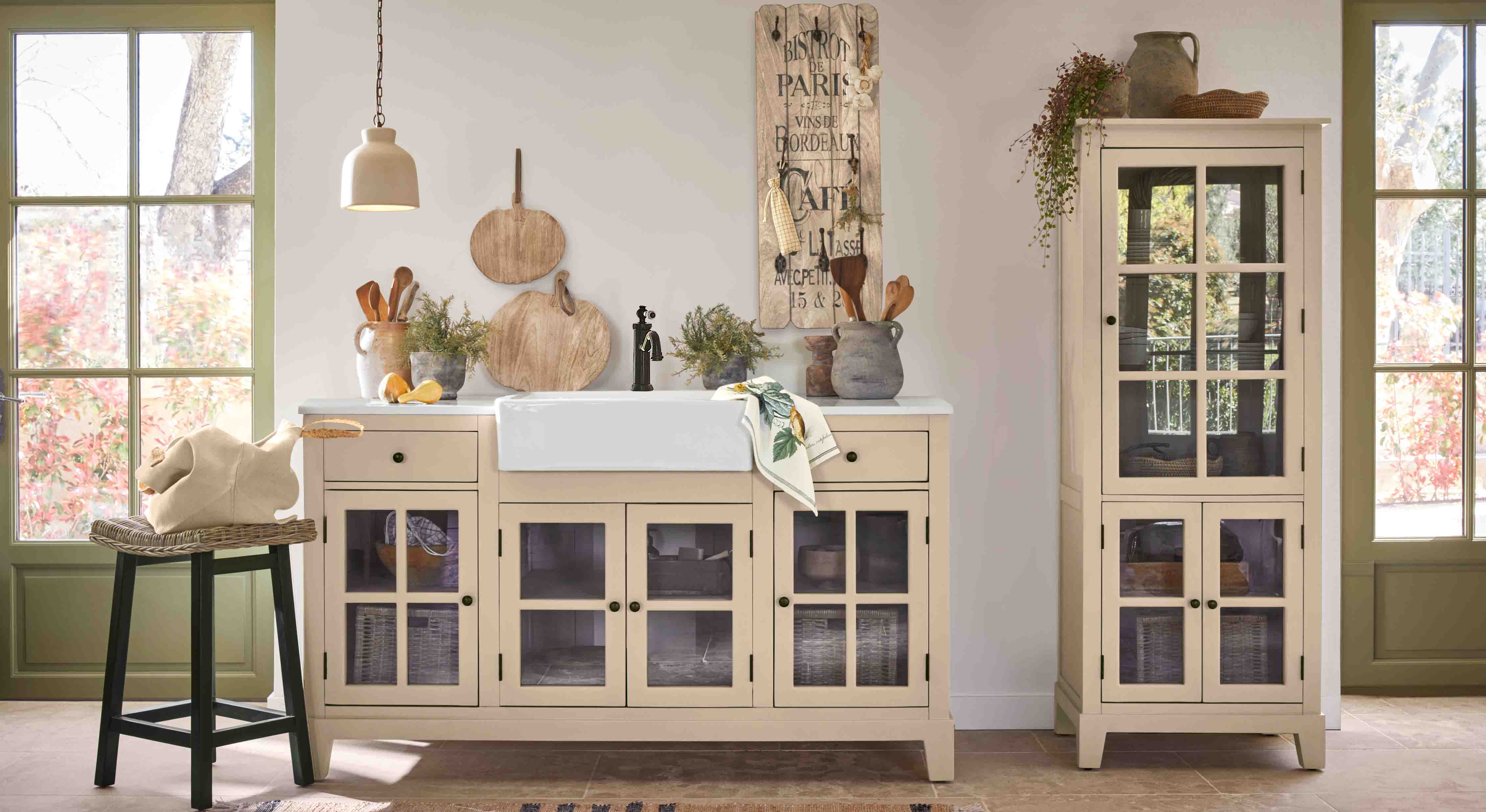 Kuchnia w stylu rustykalnym z beżowymi szafkami z szybą i białym ceramicznym zlewem, dekoracje drewniane i rośliny