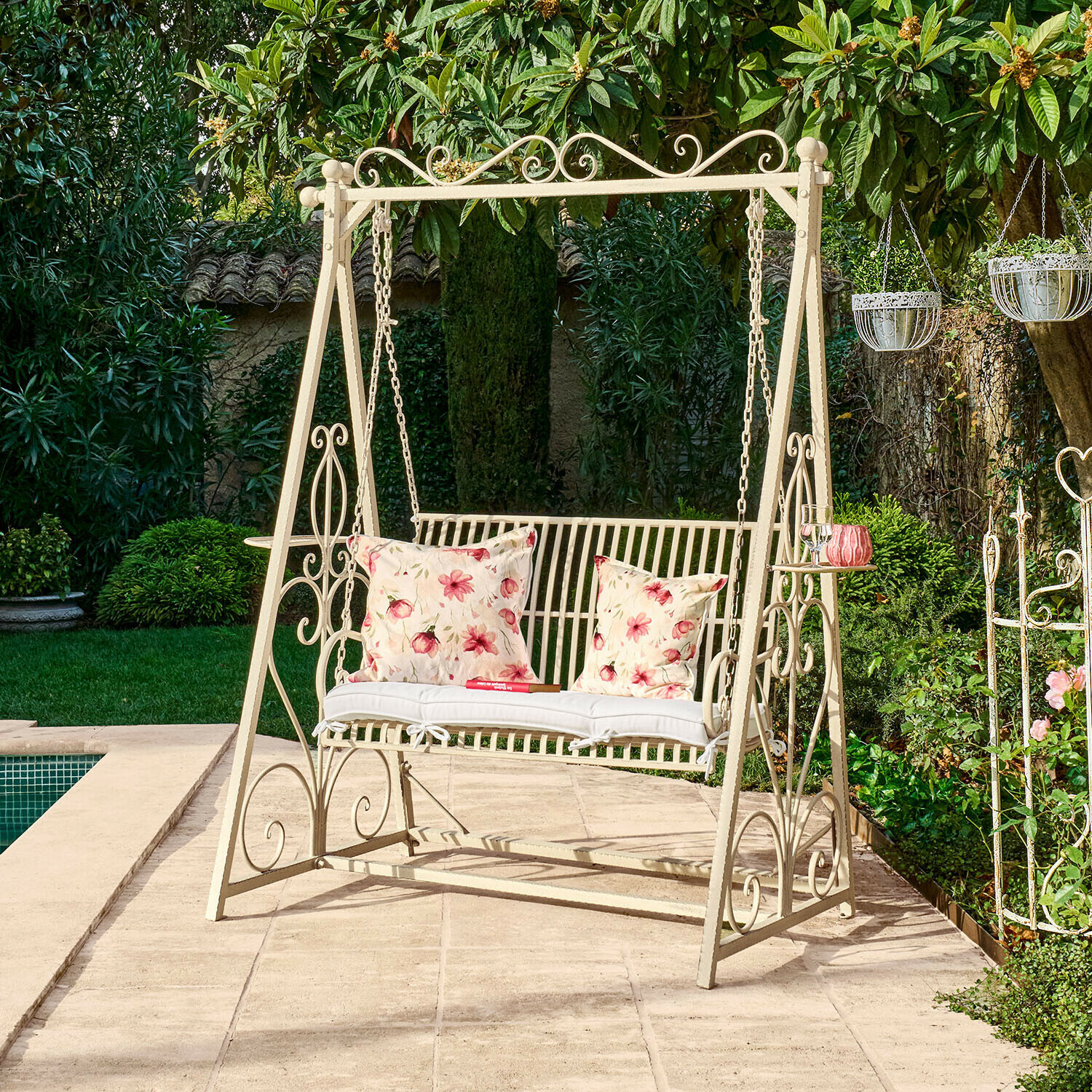 Metalowa huśtawka ogrodowa w kremowym kolorze z poduszkami w kwiatowy wzór na tarasie przy basenie