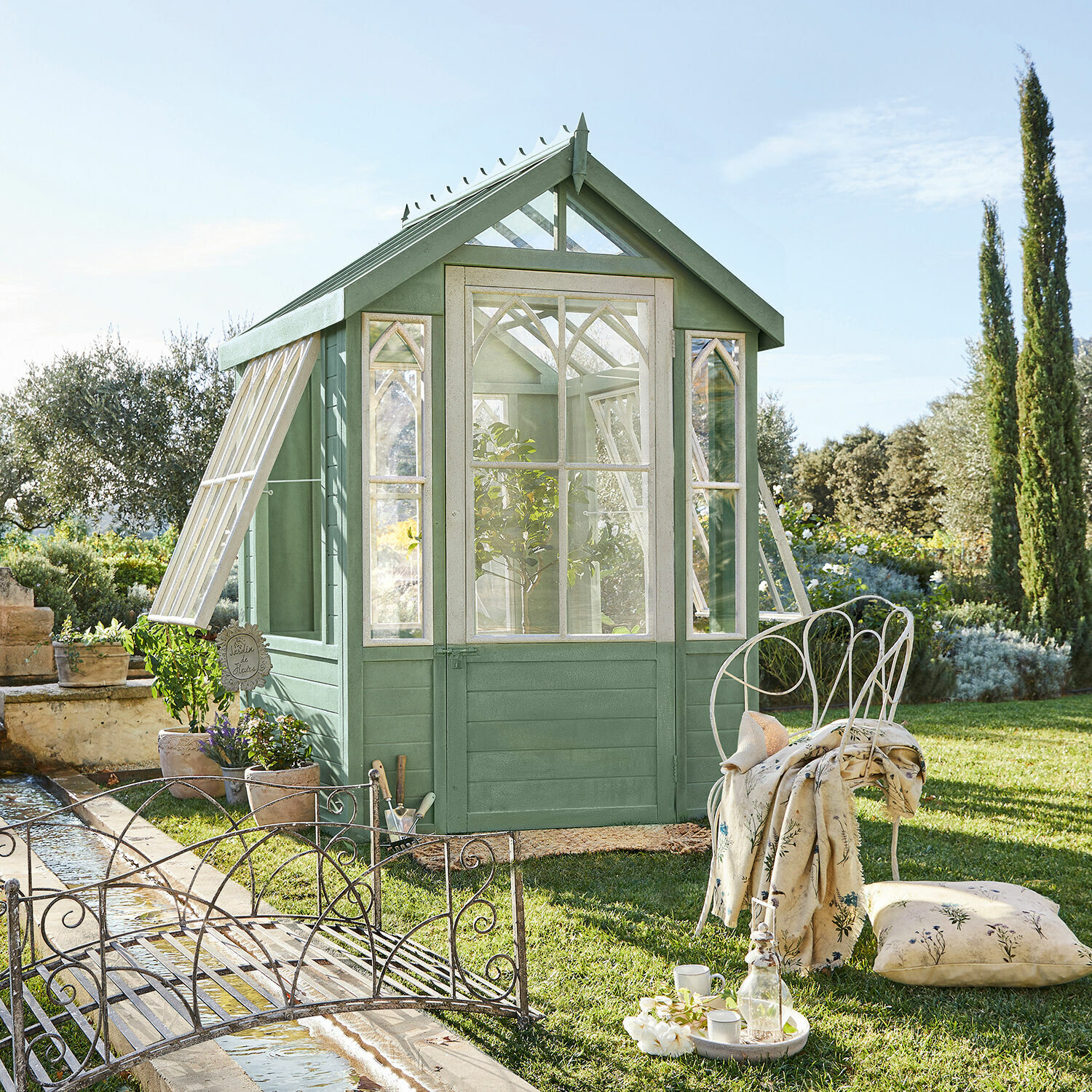 Zielona drewniana szklarnia ogrodowa z białymi oknami i metalowym meblem na trawniku