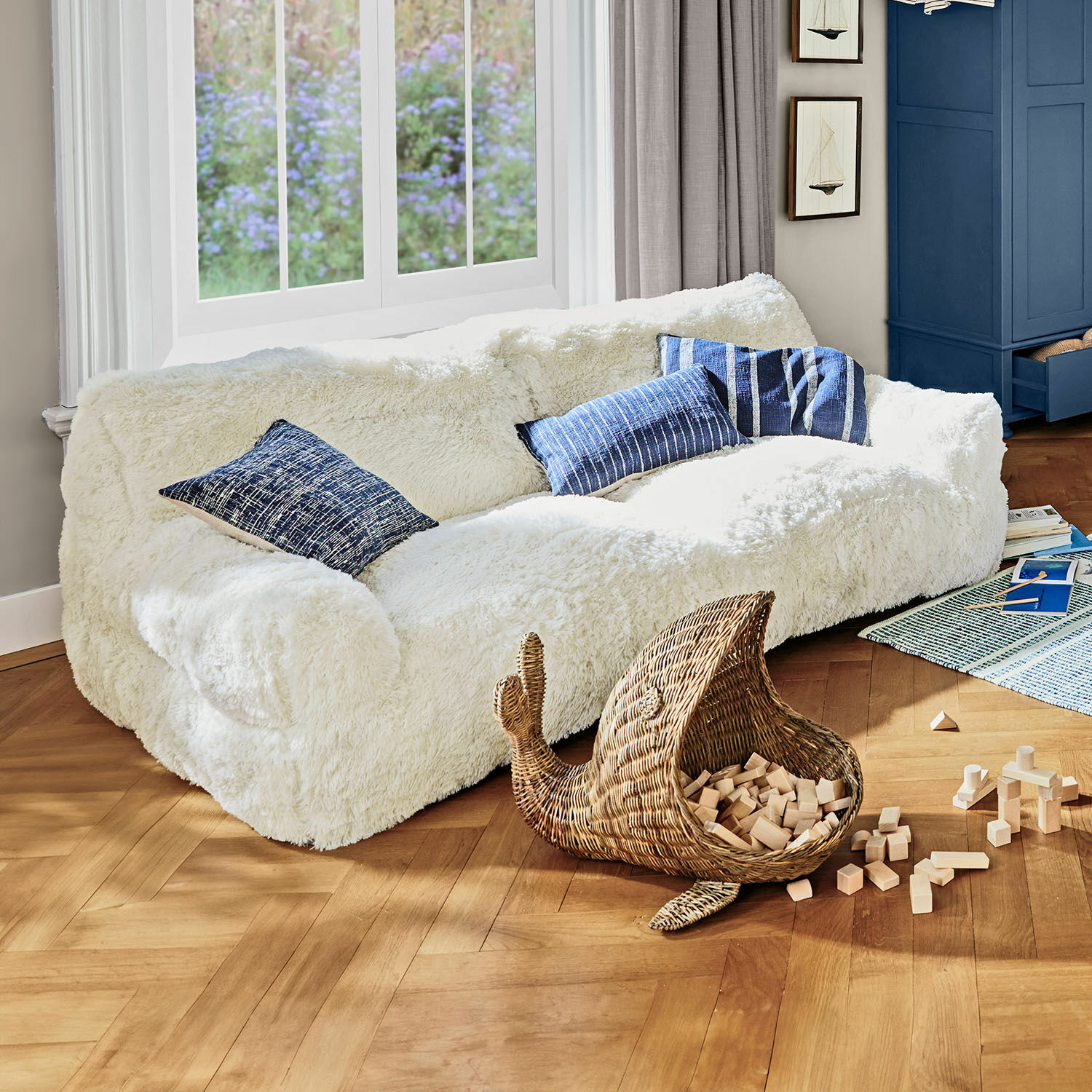 Sofa dla dzieci Cerveteri Biały, miękki, futrzany narożnik z niebieskimi poduszkami w jasnym salonie z drewnianą podłogą