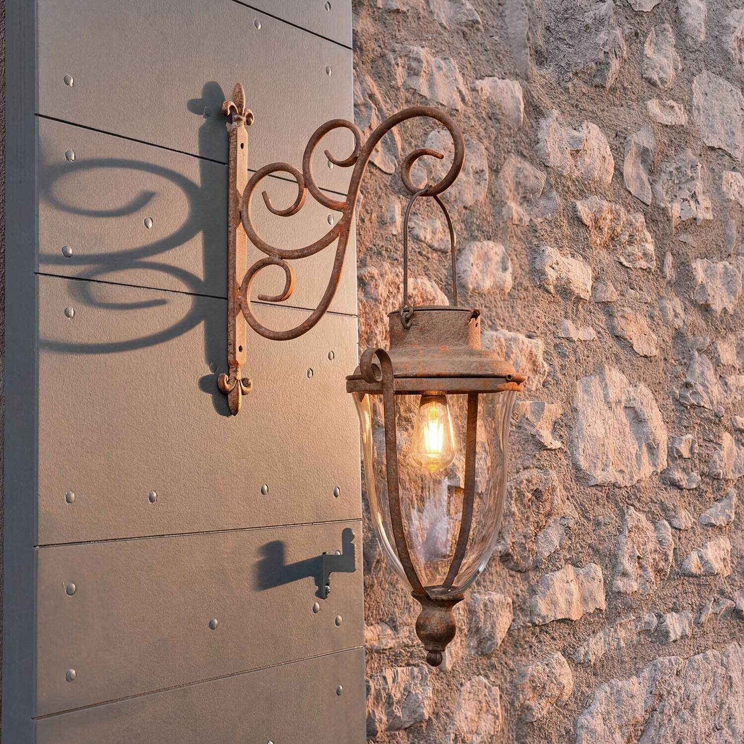 Lampion solarny Volanaïs Rustykalna zewnętrzna lampa ścienna z metalu w kolorze rdzy przy kamiennej ścianie oświetlająca wejście