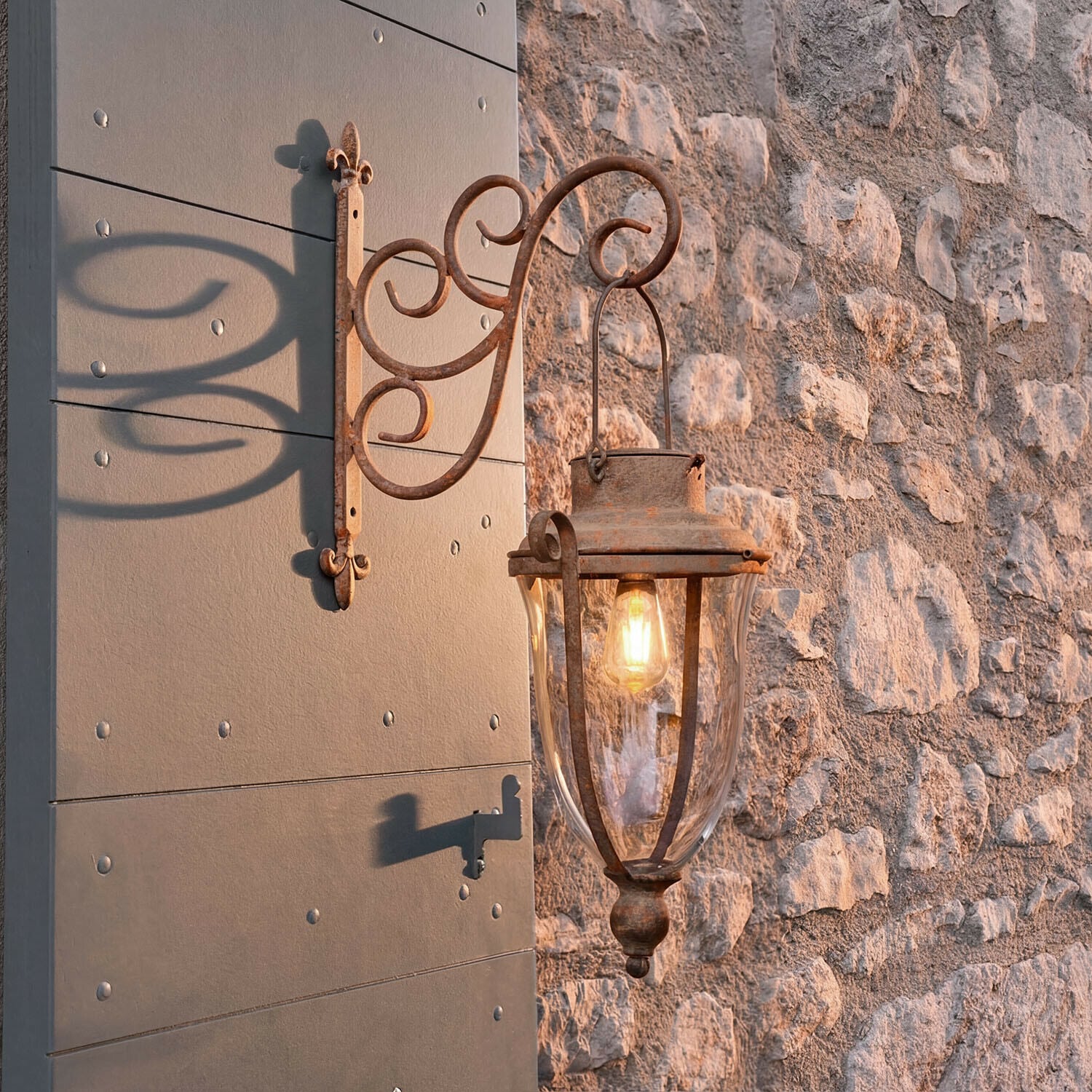 Lampion solarny Volanaïs Rustykalna zewnętrzna lampa ścienna z metalu w kolorze rdzy przy kamiennej ścianie oświetlająca wejście