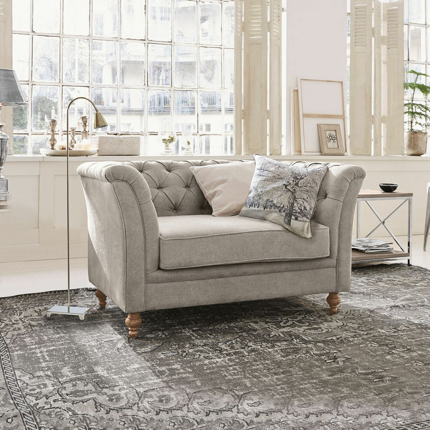 Sofa Clères Szara tapicerowana sofa z drewnianymi nogami na wzorzystym dywanie w jasnym salonie